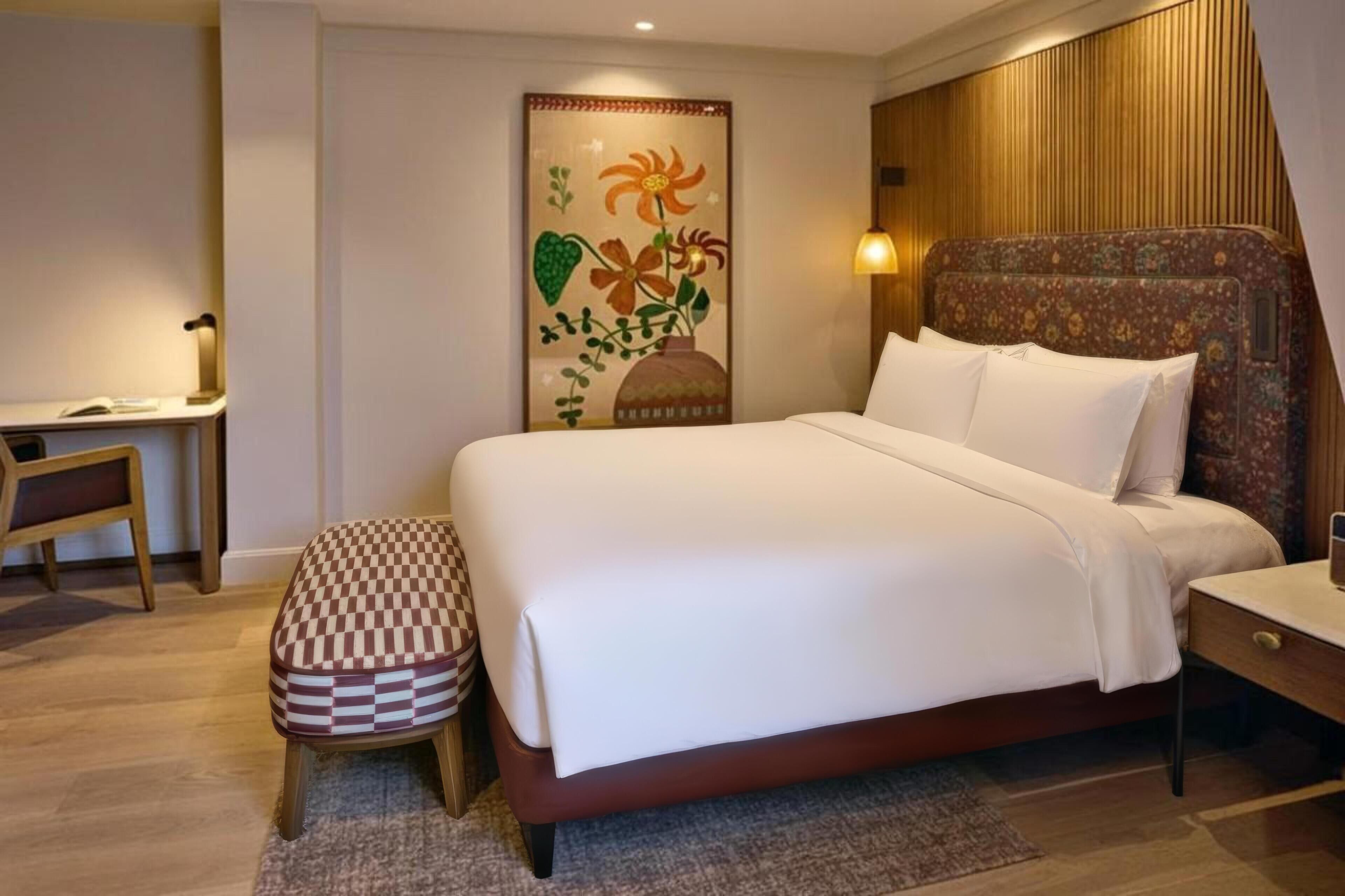 A bedroom with a bed, a desk, a chair, a bench, and a wall-mounted lamp.