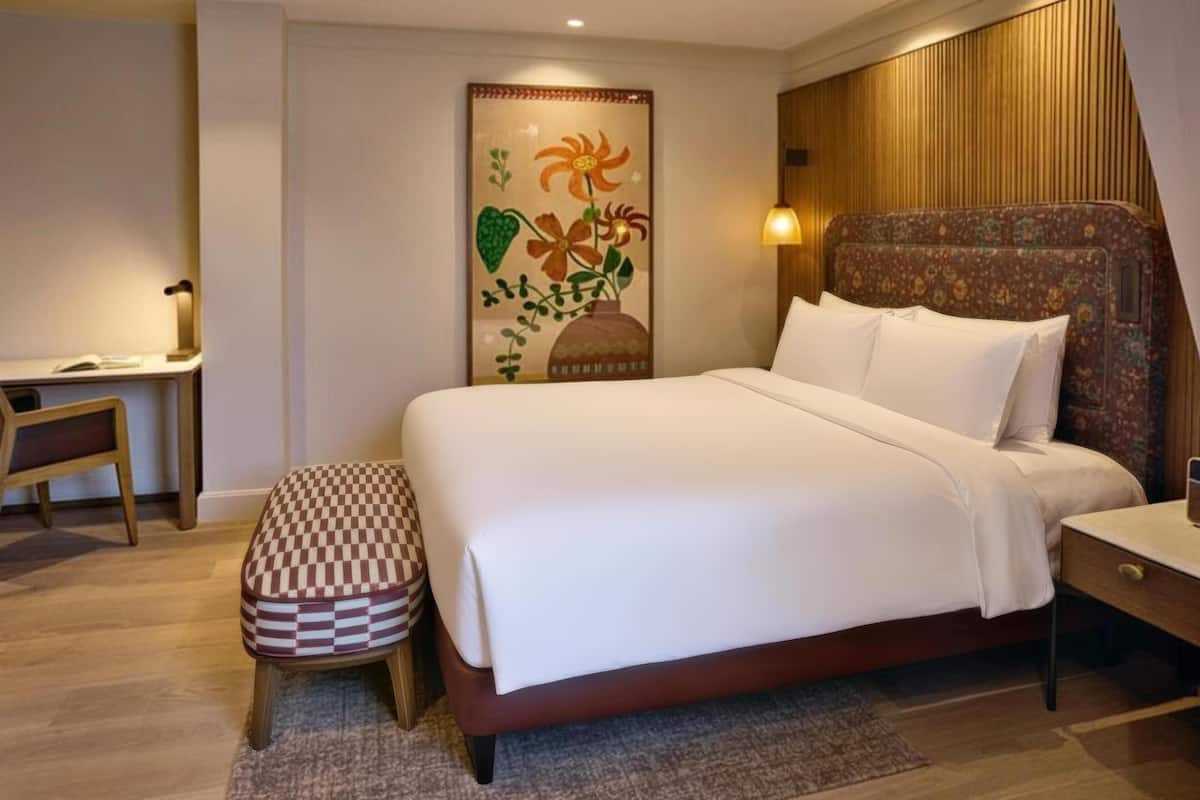 A bedroom with a bed, a desk, a chair, a bench, and a wall-mounted lamp.