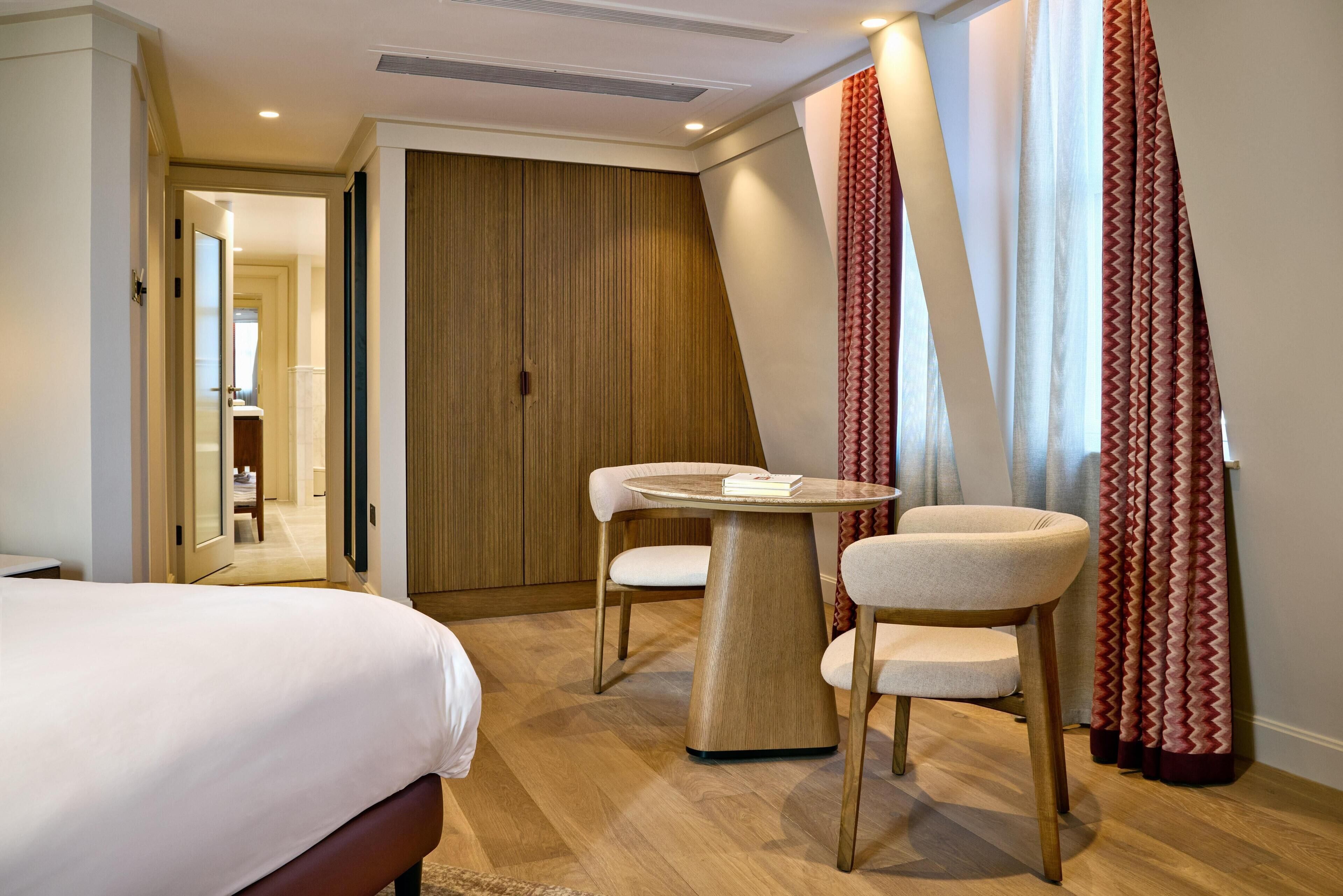 A hotel room with a bed, a round table, two chairs, a wooden wardrobe, and a view of the outside through curtains.
