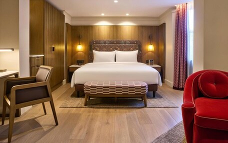 A hotel room with a large bed, bedside tables, a desk, a chair, and a red armchair.