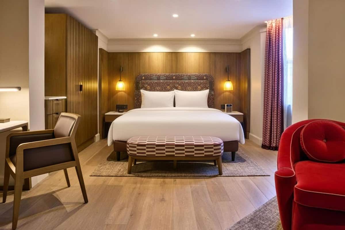A hotel room with a large bed, bedside tables, a desk, a chair, and a red armchair.