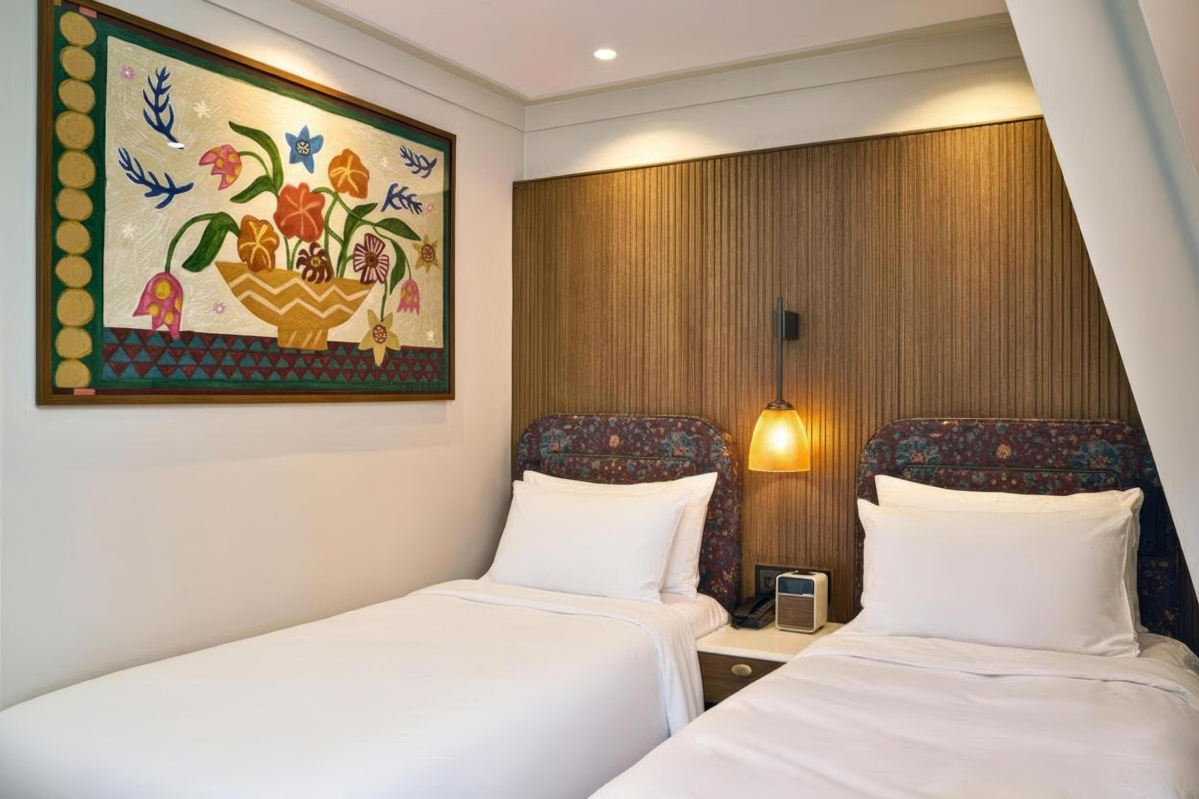 A hotel room with two beds, a wooden headboard, a wall-mounted painting, and a bedside lamp.
