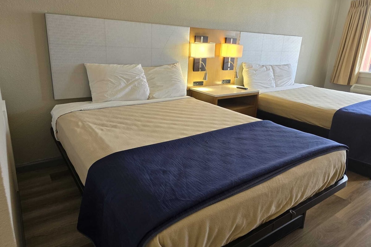 Habitación de hotel con dos camas, cada una con cabecera, mesita de noche con lámparas y ventana con cortinas.