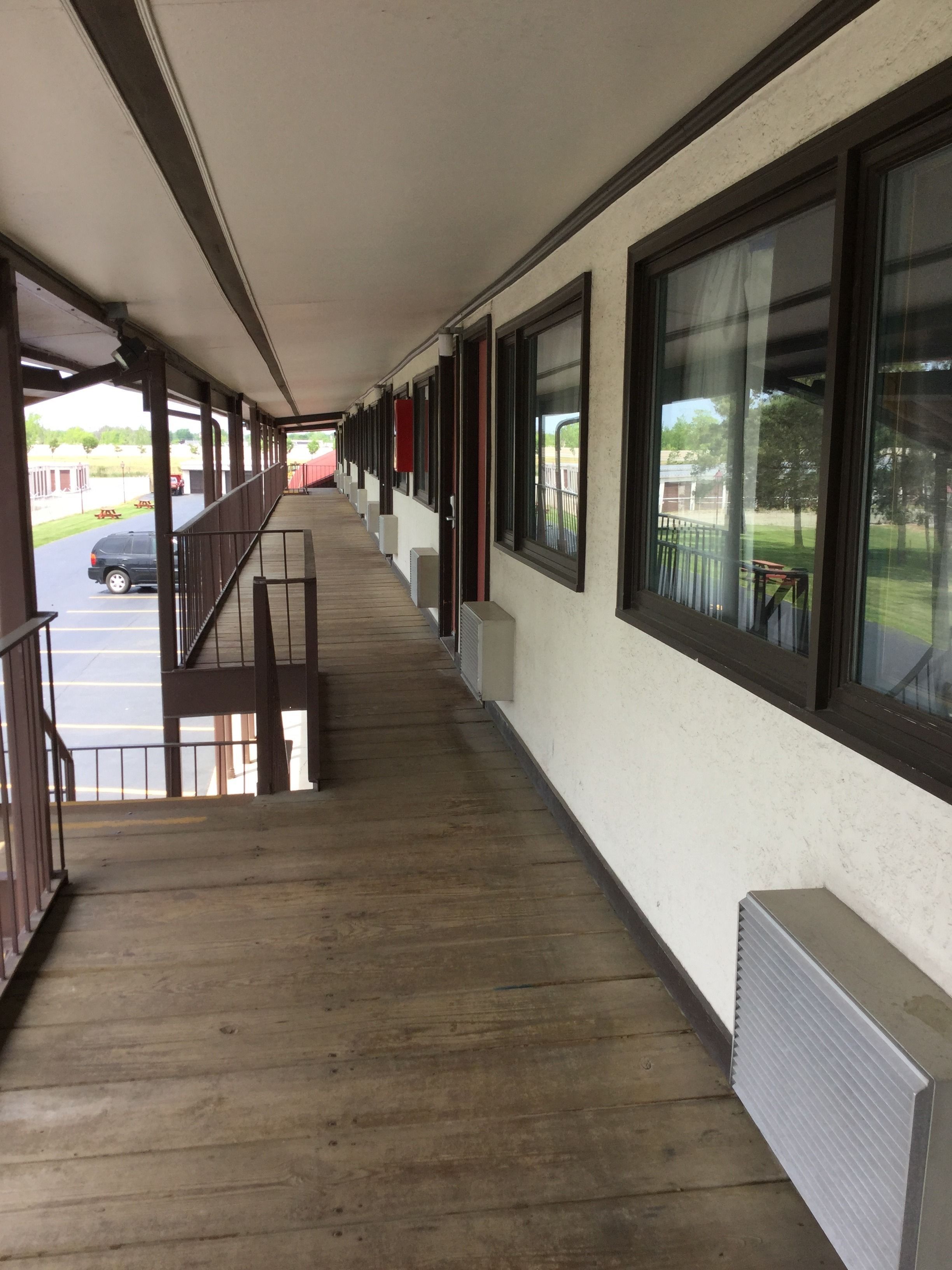 Un couloir couvert doté d’un plancher en bois, de grandes fenêtres et offrant une vue sur un parking et des espaces verts à l’extérieur.
