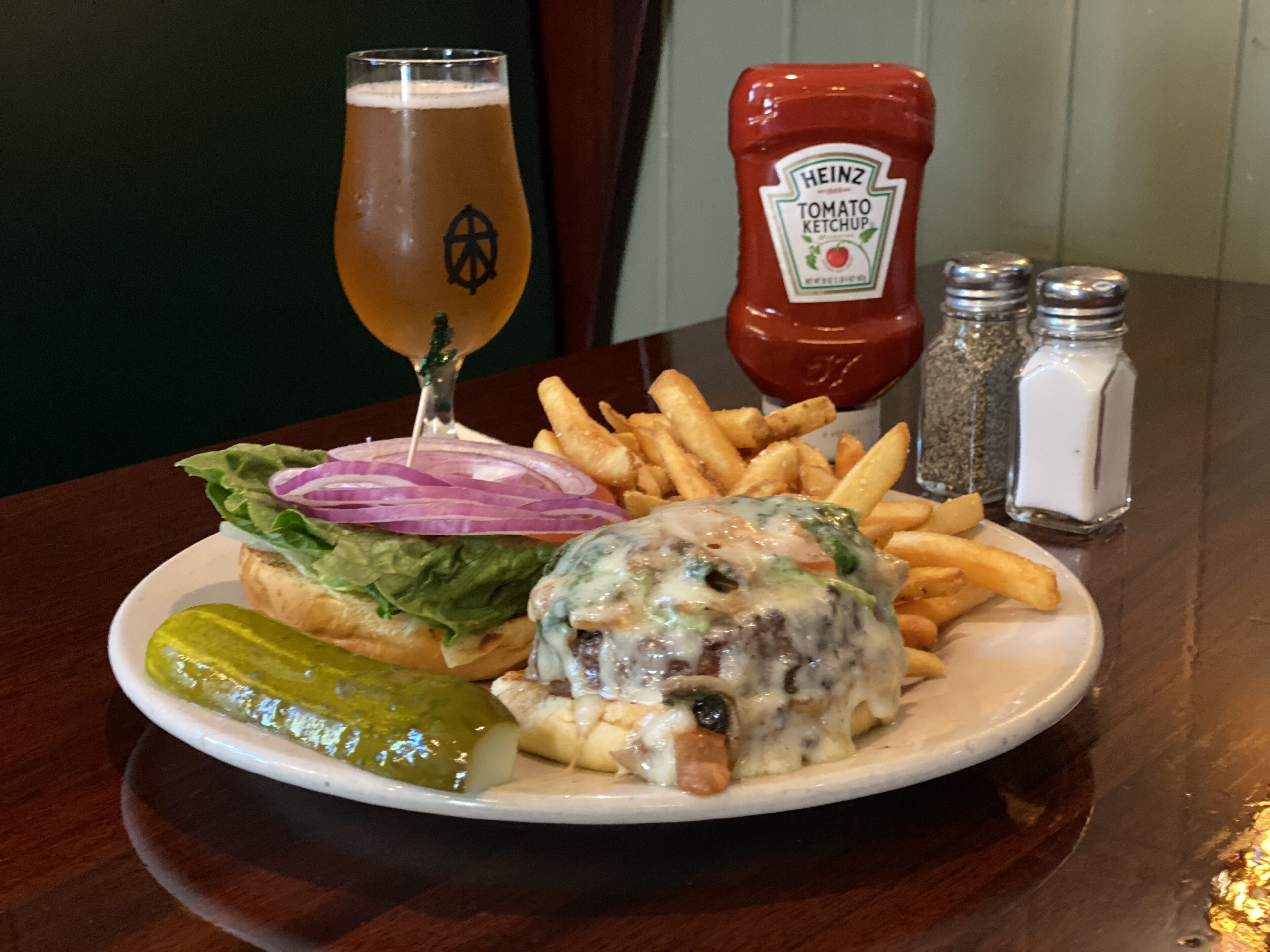 A plate with a burger, fries, and a pickle, a glass of beer, and condiments on a table.