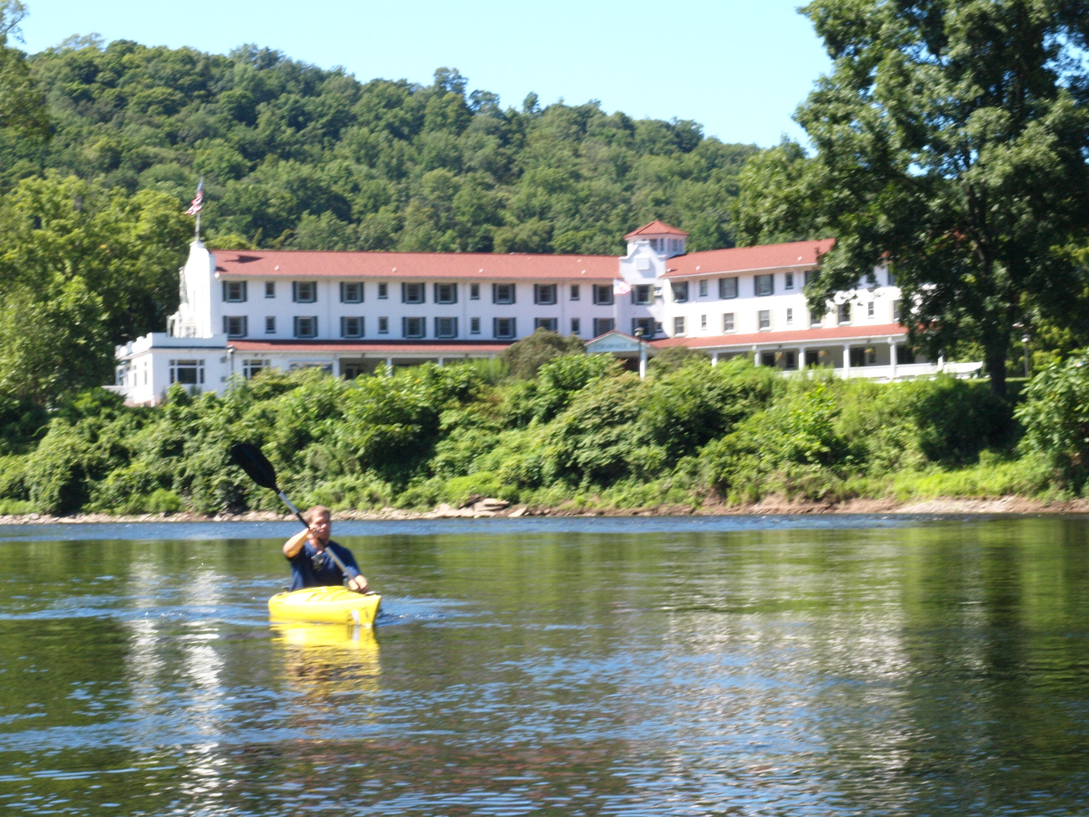 Una persona en un kayak amarillo sobre un lago tranquilo, con un edificio grande de techos rojos al fondo.