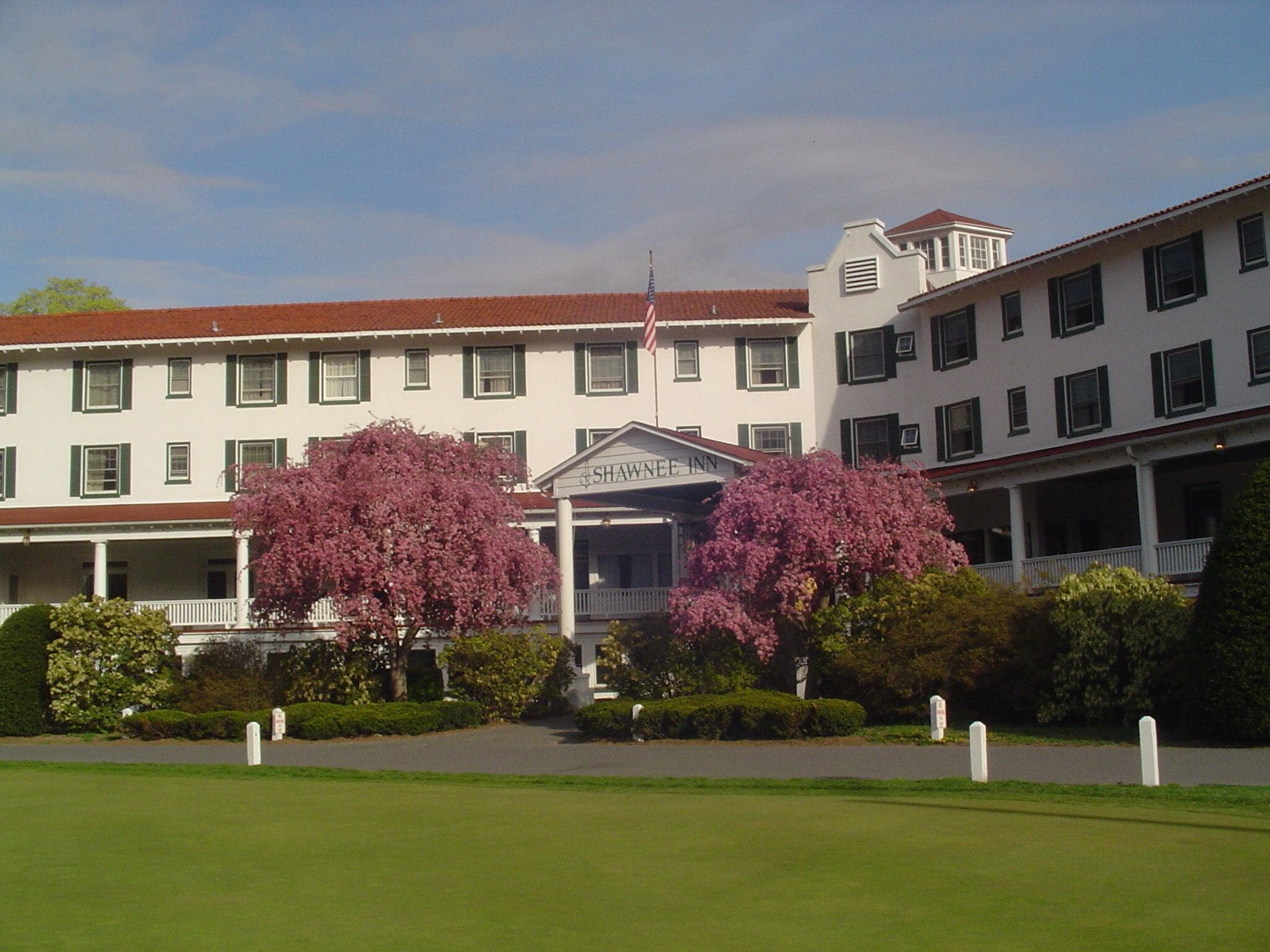 Un edificio grande y blanco con techo rojo, un cartel que dice "Shawnee Inn" y dos árboles grandes con flores rosadas al frente.