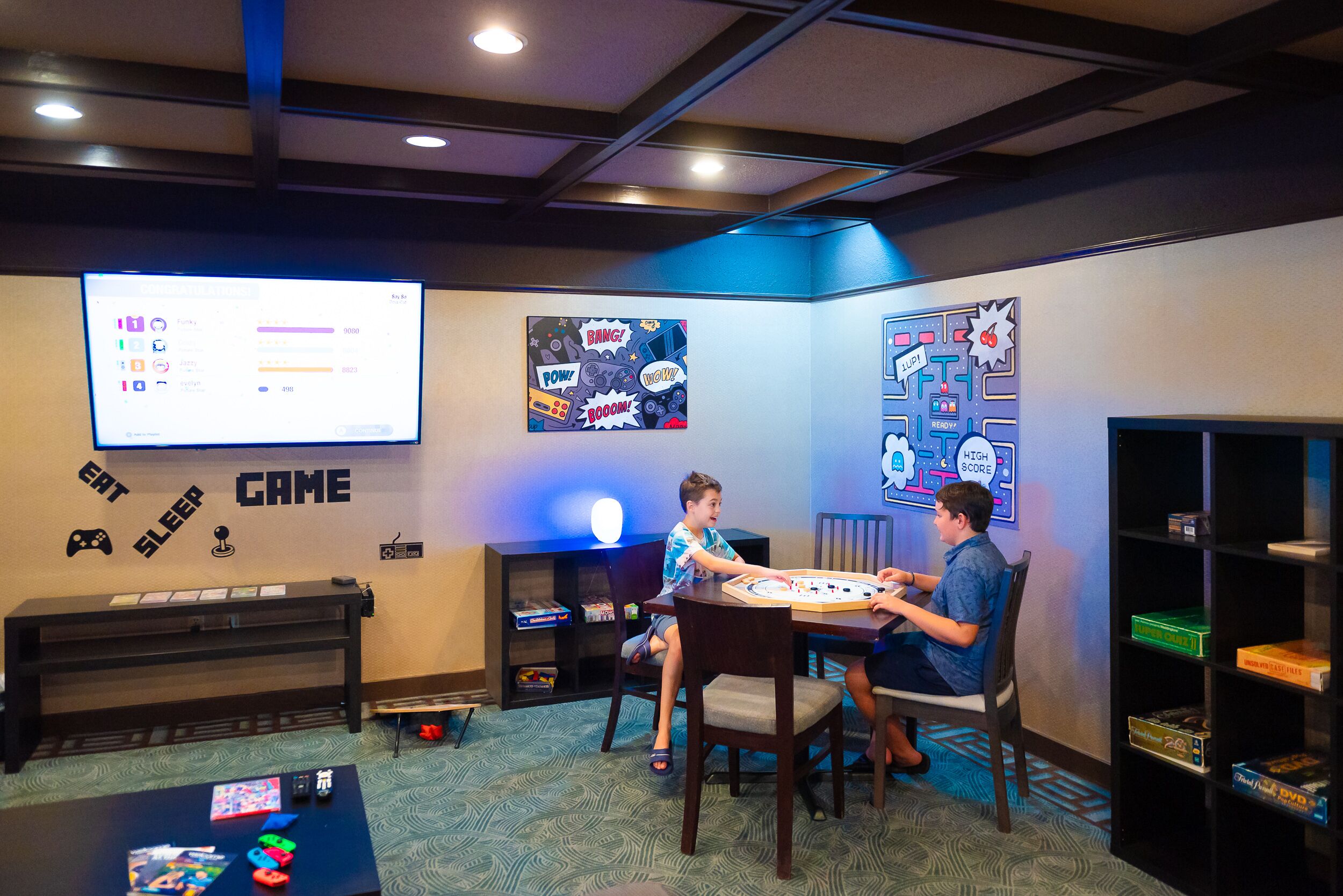 A gaming room with a large screen displaying game stats, a table with board games, and a bookshelf with board games.