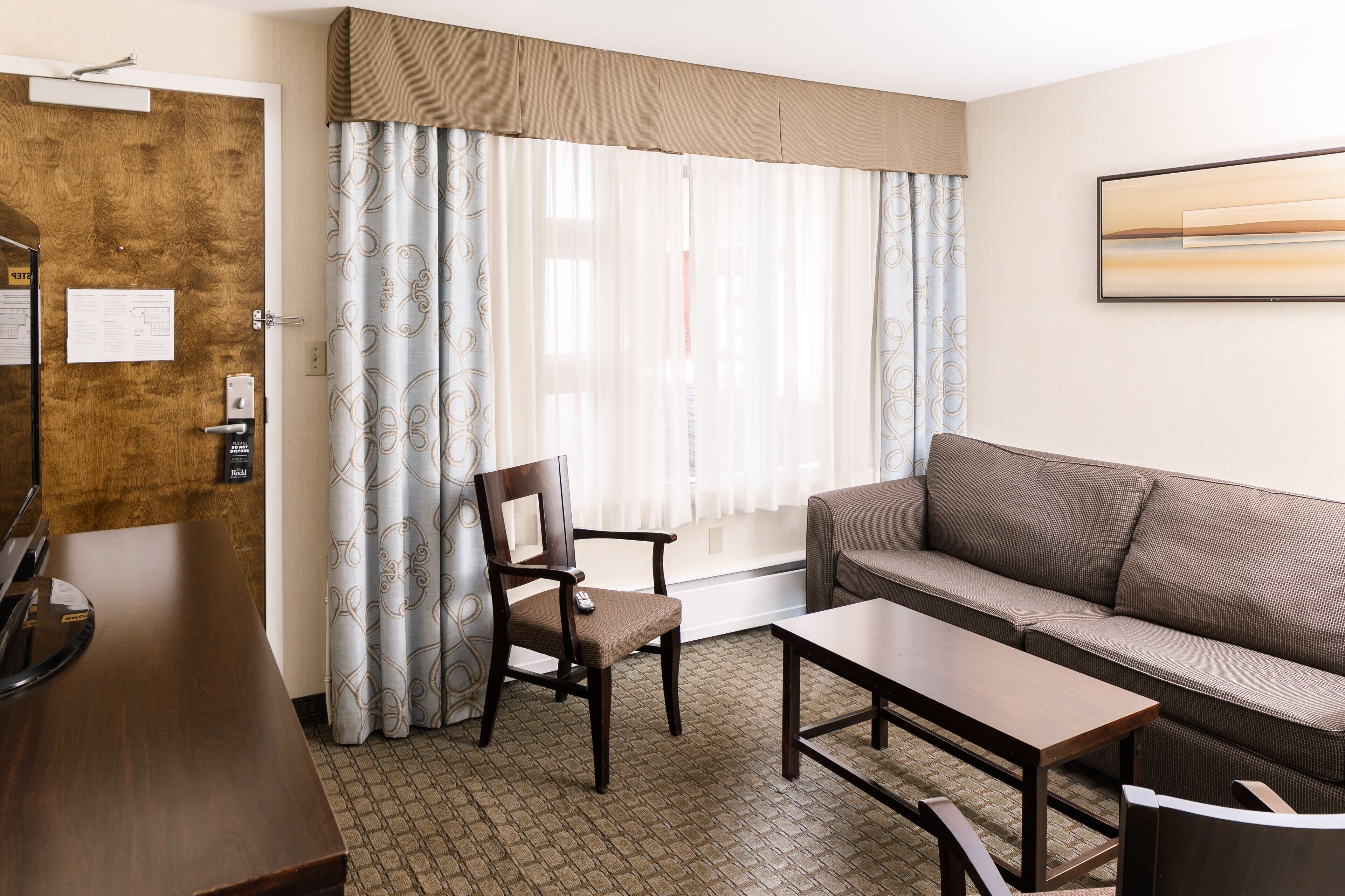 A room with a wooden door, a desk with a telephone, a chair, a sofa, and a window with curtains.