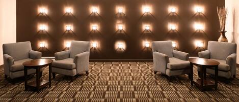 Un vestíbulo de hotel con alfombra estampada, cuatro sillones, dos mesas redondas pequeñas y una pared con varias luces empotradas.