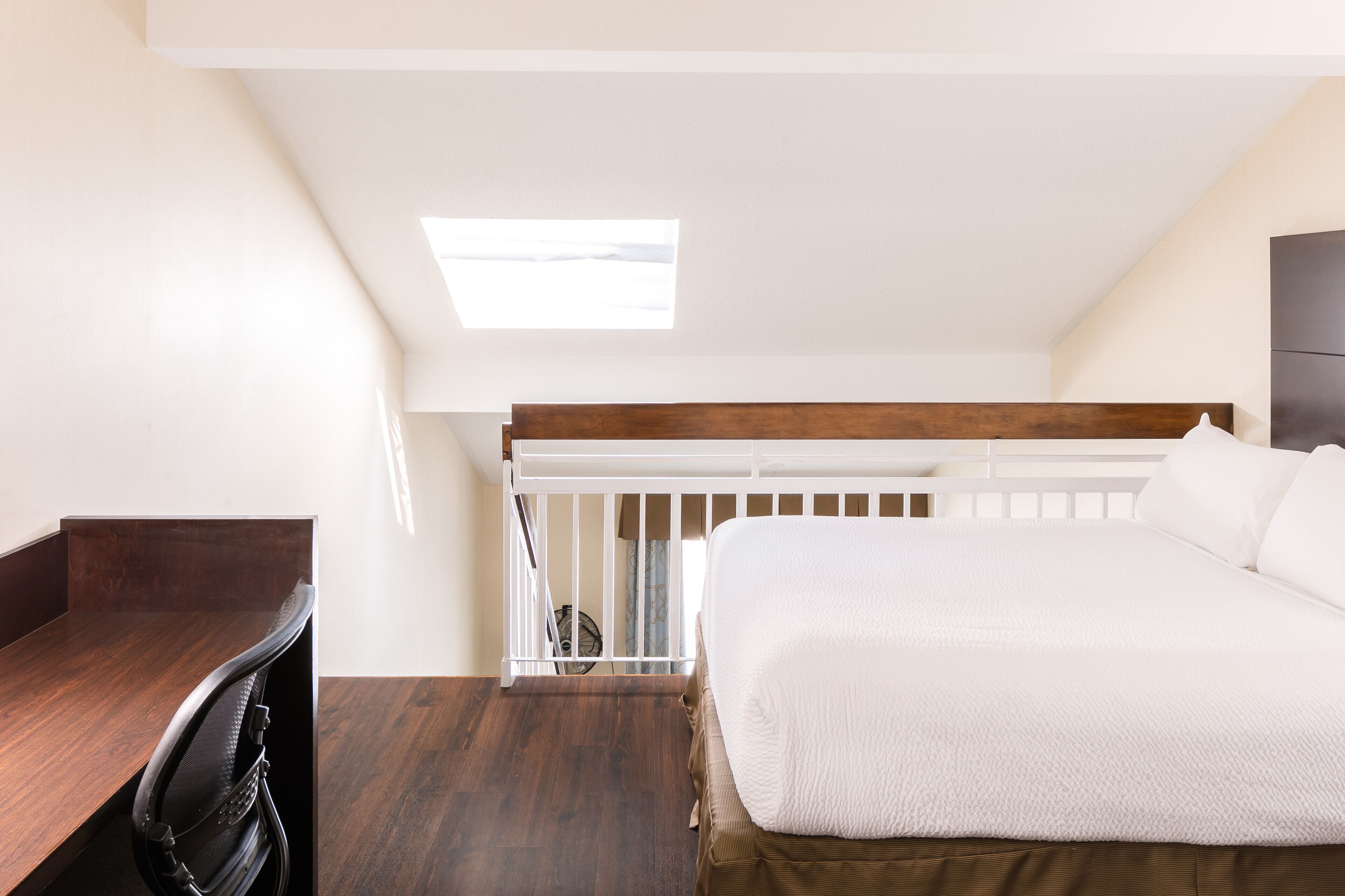 Ein Schlafzimmer mit schrägen Wänden, einem Bett mit weißer Bettwäsche, einem Schreibtisch mit Stuhl und einem kleinen Fenster.