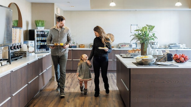 A modern kitchen with wooden cabinets, a central island with food items, and a refrigerator with a plant on top. - Rodd Royalty (Charlottetown)