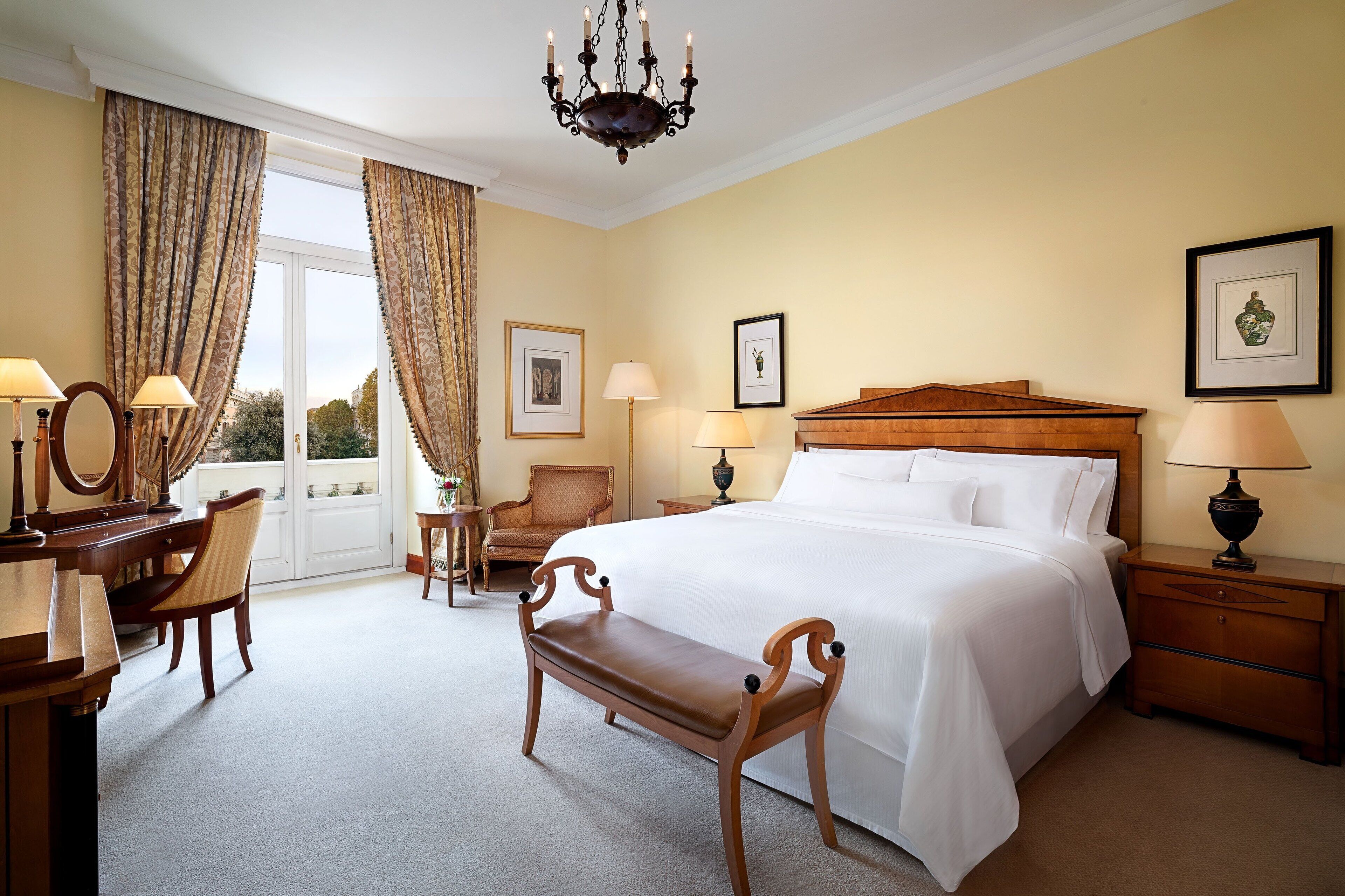 A hotel room with a large bed, a wooden bench, a desk with a mirror, two chairs, a nightstand with a lamp, and a chandelier.