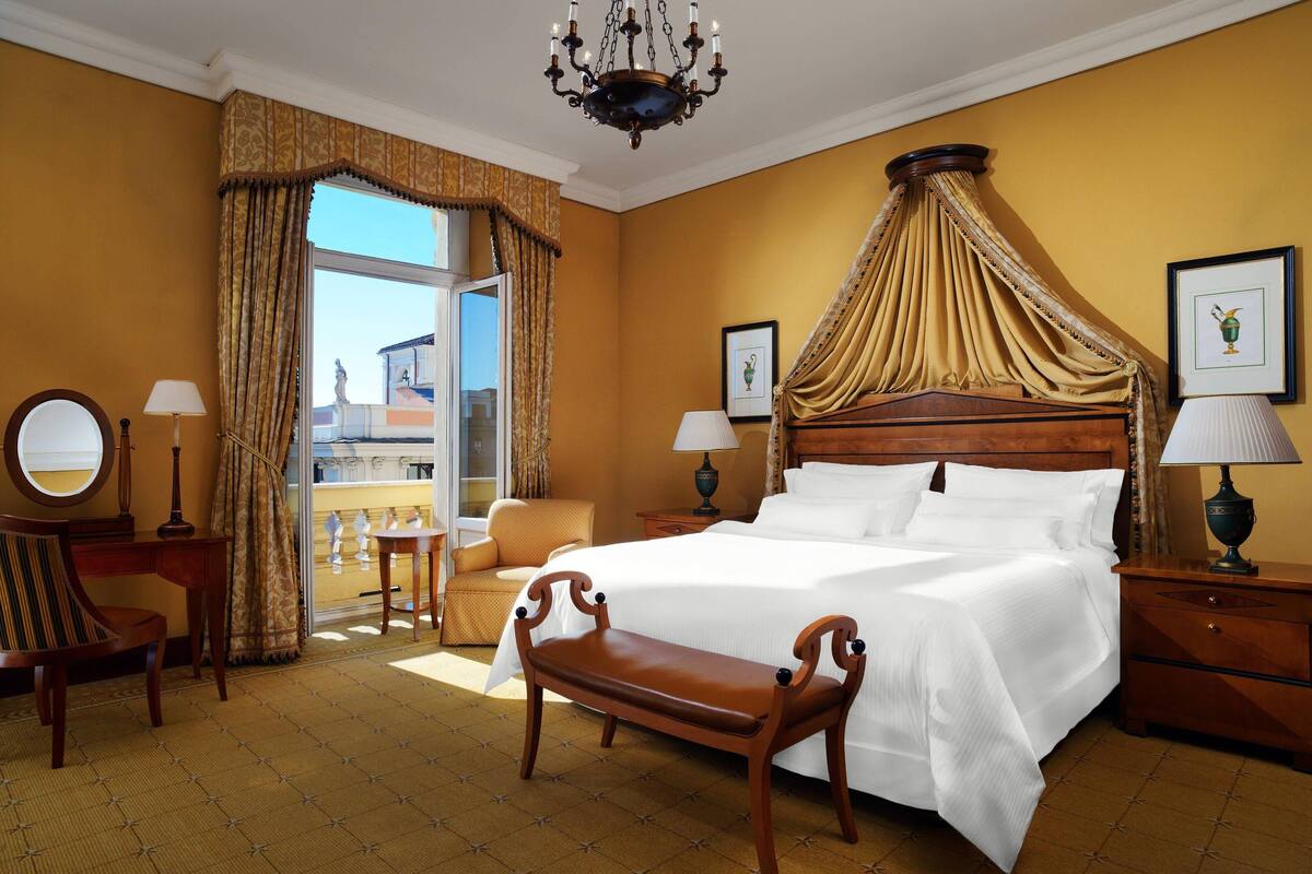 A hotel room with a large bed, a bench, a desk, a chair, a nightstand, and a chandelier.