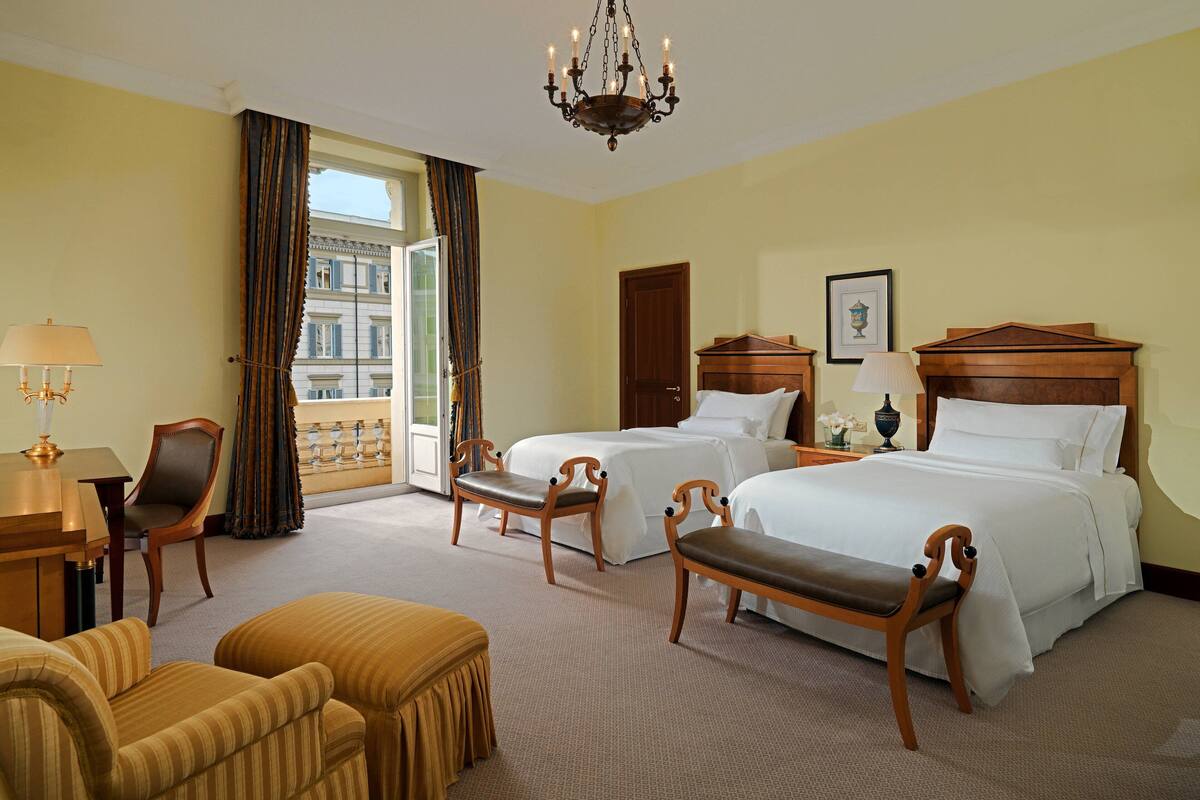 A hotel room with two beds, a sitting area with a bench, a desk with a lamp, and a view of a building through a window.