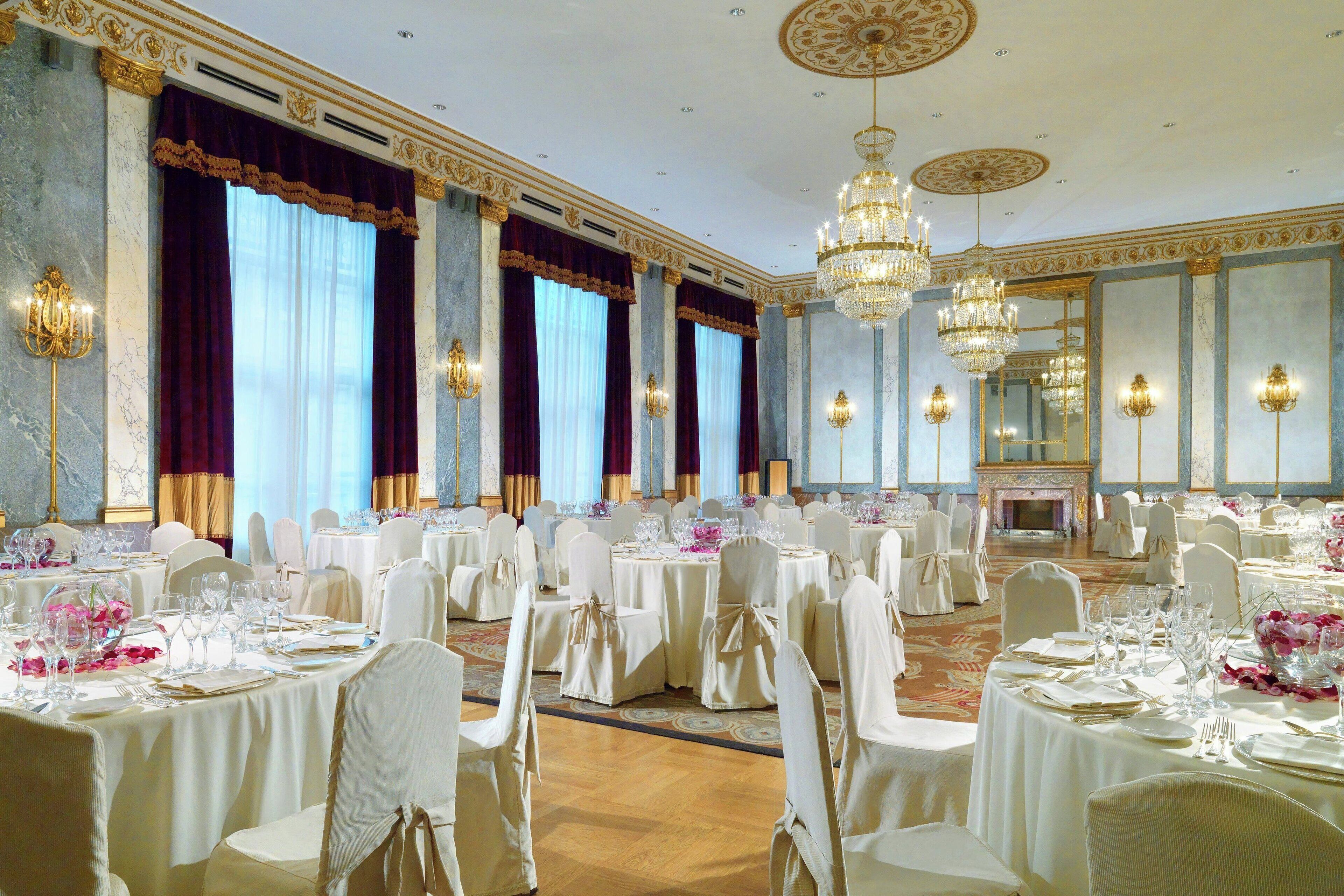 A banquet hall with round tables, white chairs, chandeliers, and a fireplace.