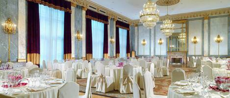 A banquet hall with round tables, white chairs, chandeliers, and a fireplace.