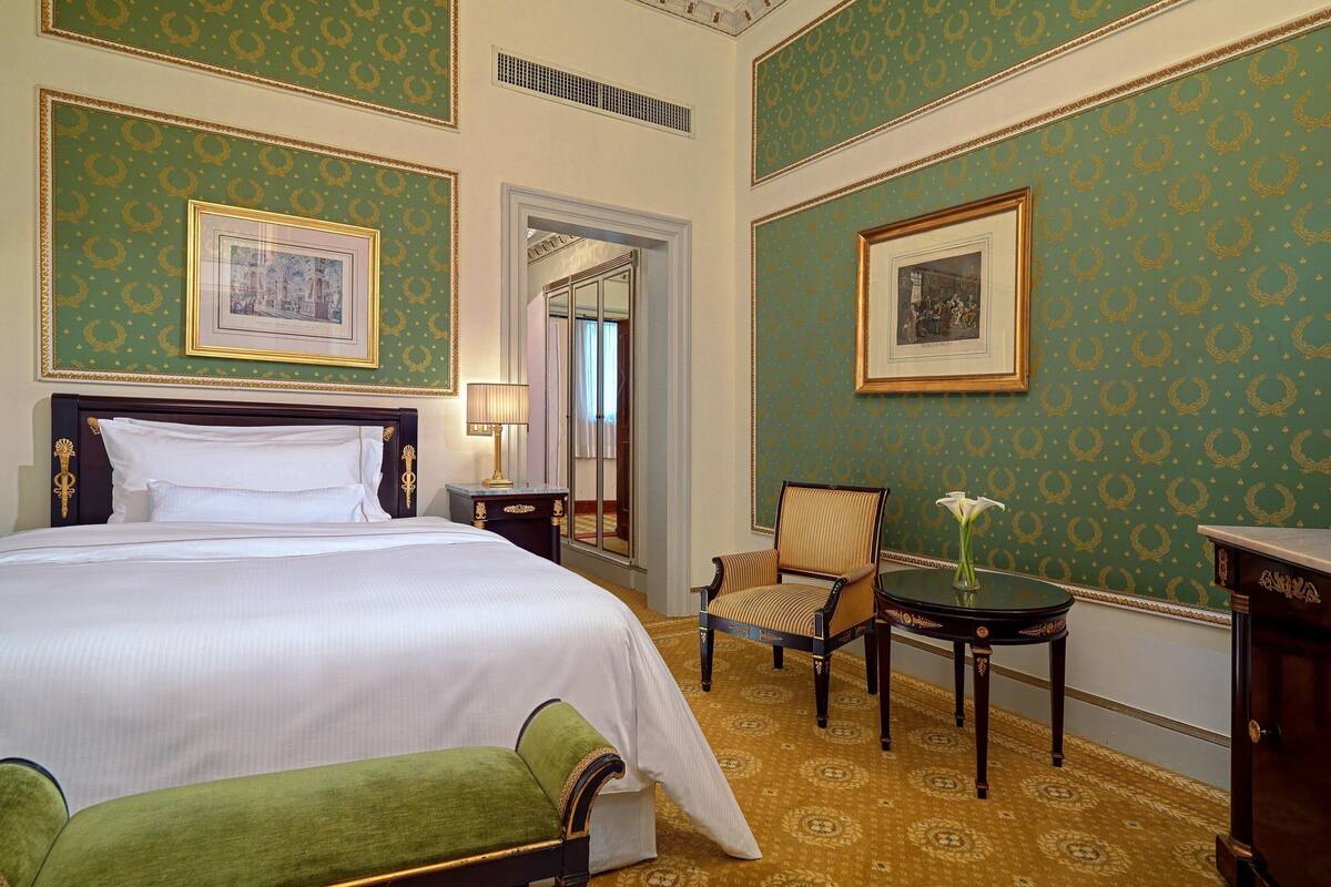 A hotel room with a large bed, a chair, a small table with a vase, and two framed pictures on the walls.