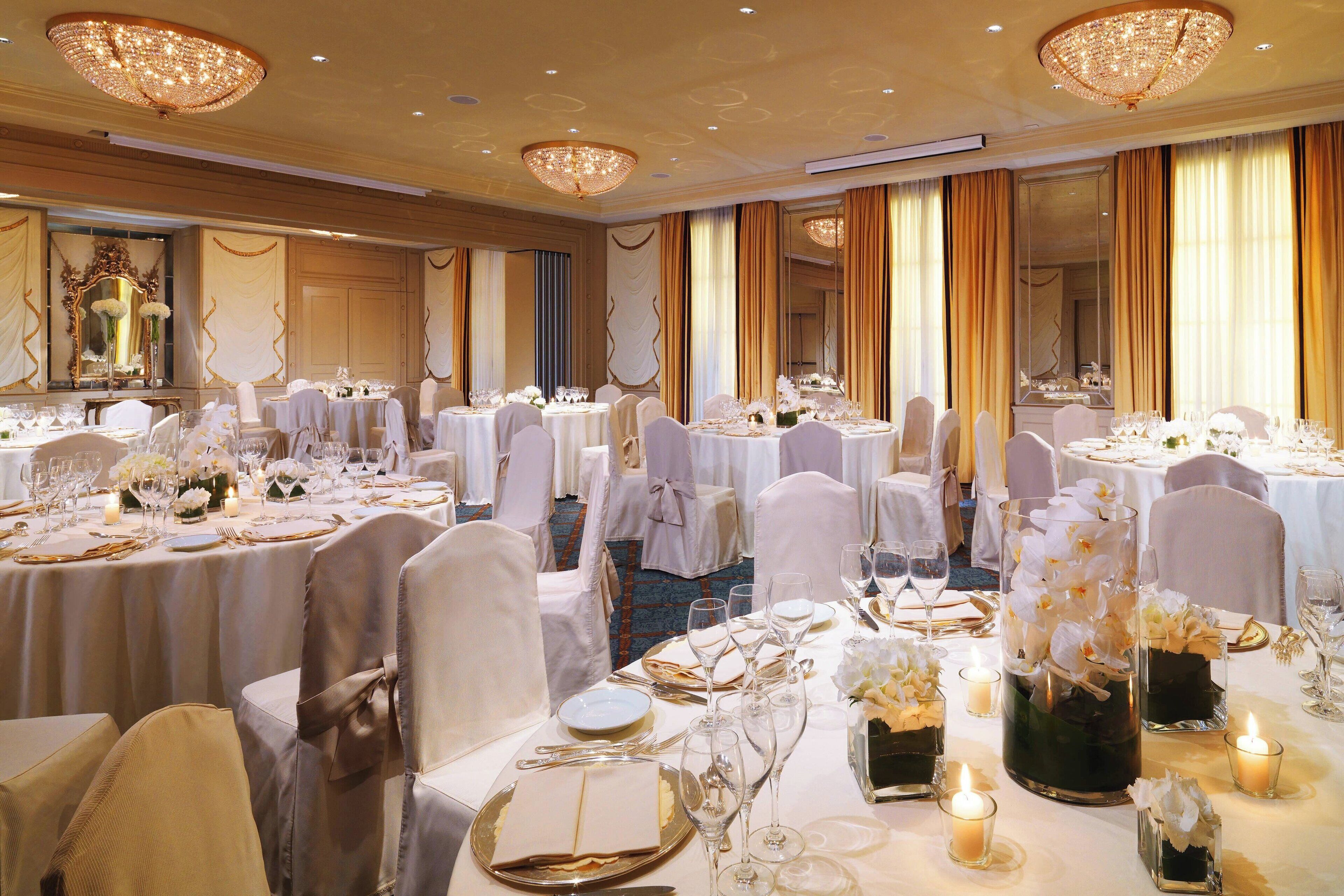 A banquet hall with round tables set for a formal event, featuring chandeliers, white tablecloths, and floral centerpieces.