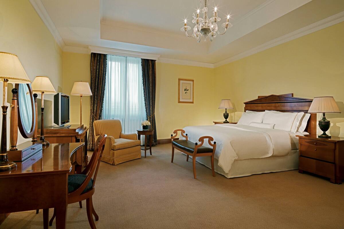 A hotel room with a bed, a desk with a chair, a bedside table with lamps, a mirror, and a chandelier.