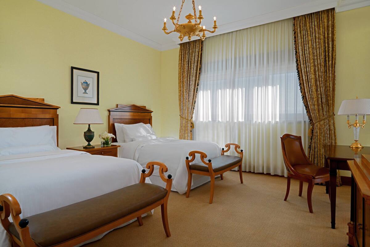 A hotel room with two beds, a wooden bench, a desk, and a chandelier.