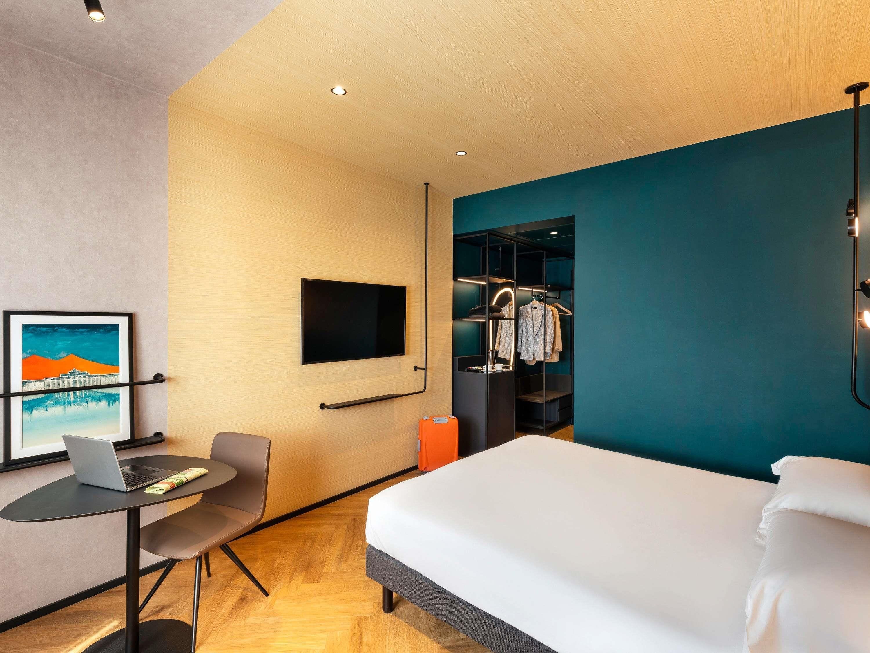 Ein modernes Hotelzimmer mit Bett, Schreibtisch mit Laptop, Fernseher, Kleiderschrank und einem Bild an der Wand.