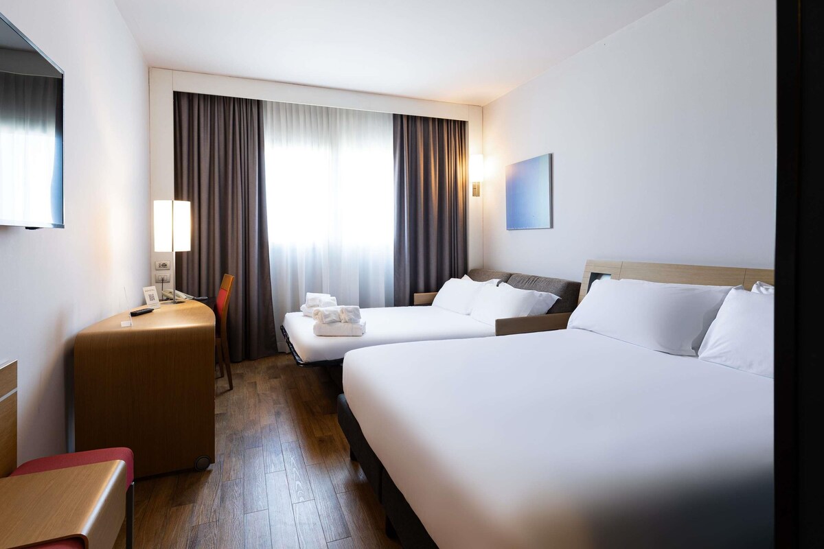 A hotel room with two beds, a desk, a chair, a TV, and a window with curtains.