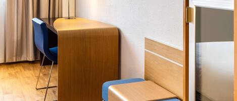 A hotel room with a wooden desk, a blue chair, a television, and a lamp.