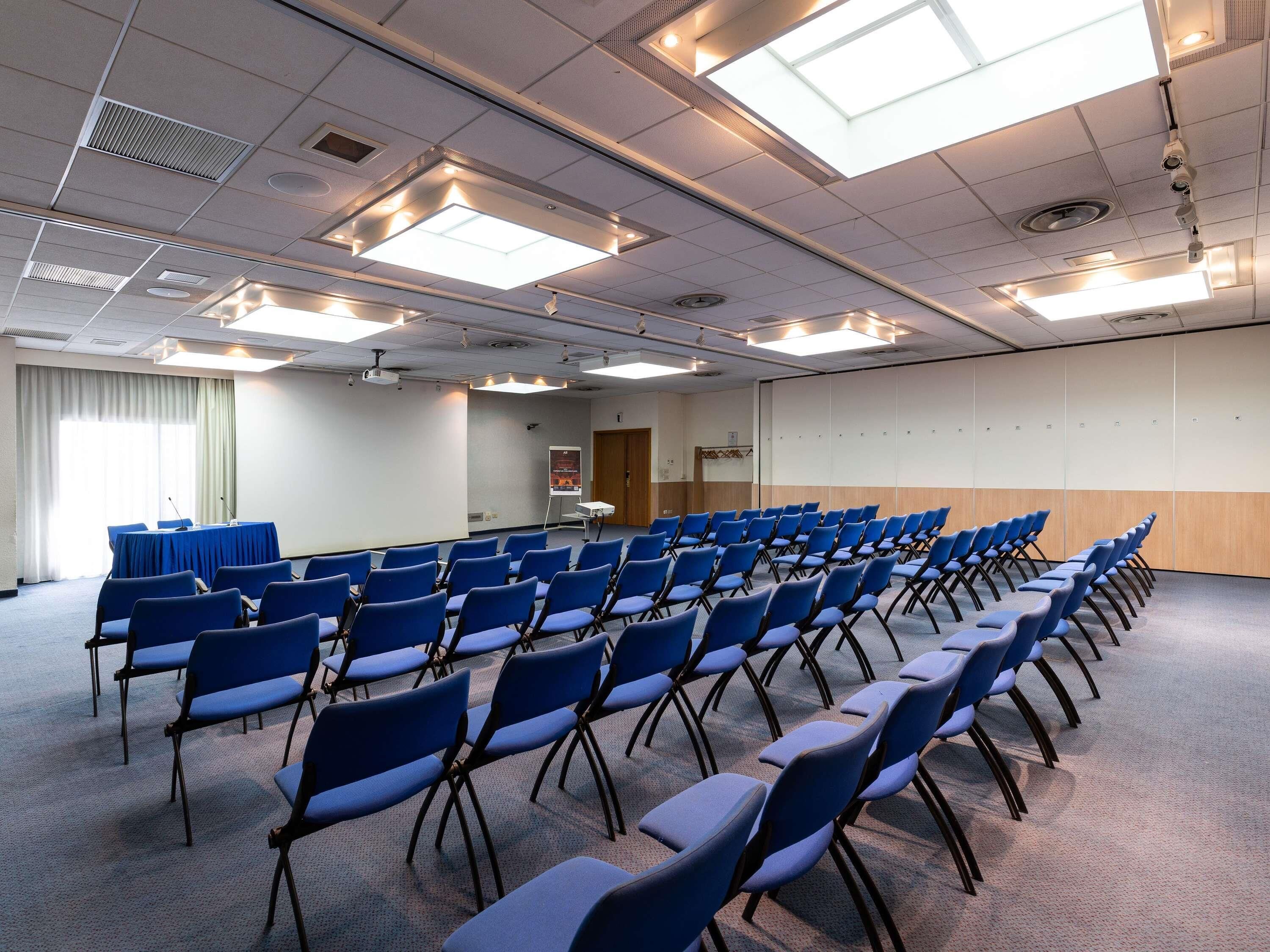 A conference room with rows of blue chairs, a presentation screen, and ceiling lights.