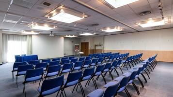 A conference room with rows of blue chairs, a presentation screen, and ceiling lights.
