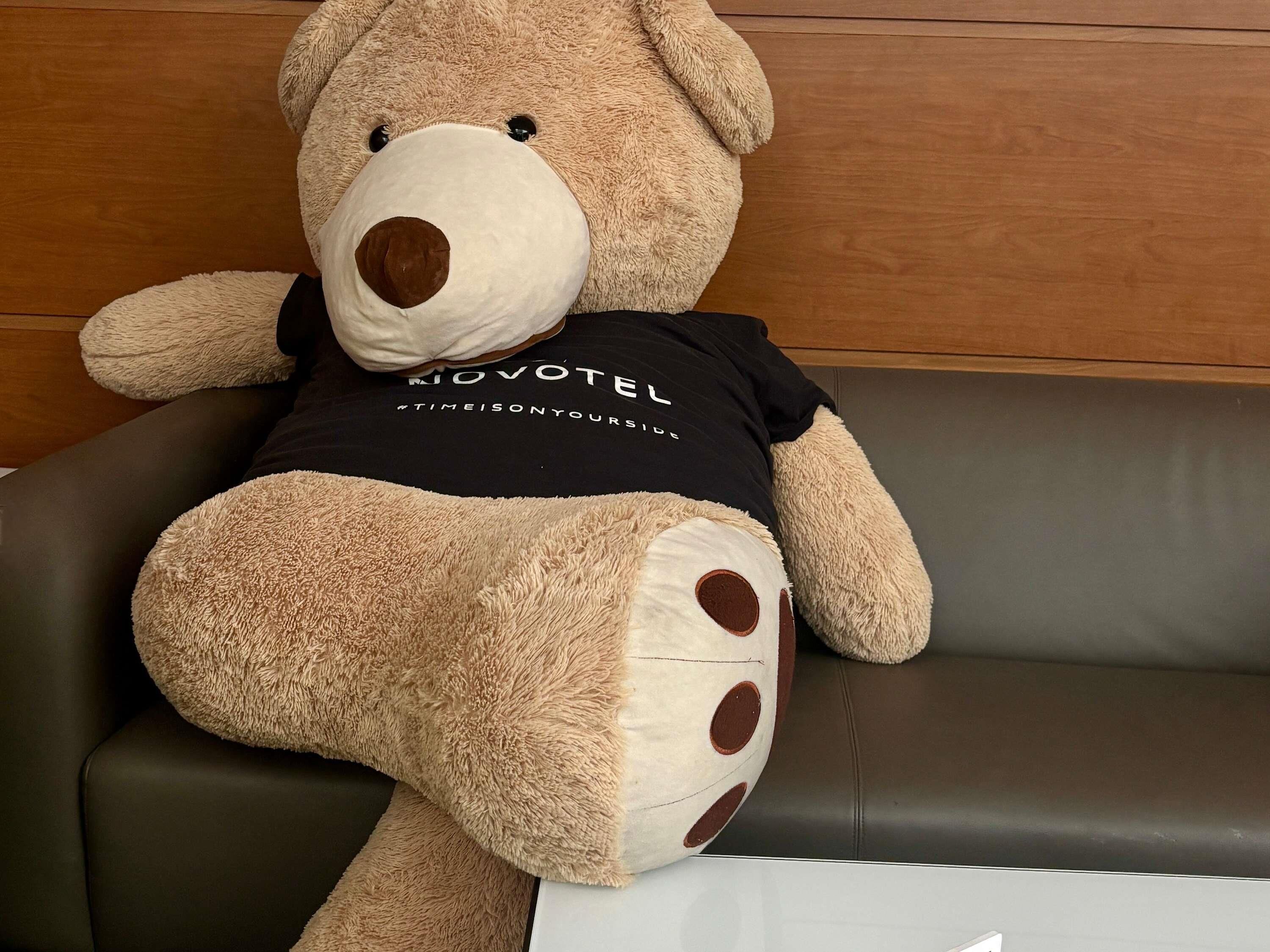 A large teddy bear sitting on a dark leather couch with a wooden panel wall in the background.