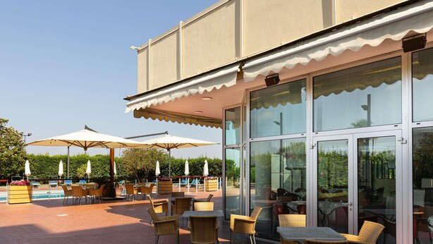An outdoor patio with wicker chairs and umbrellas, a pool in the background, and a building with large glass doors.