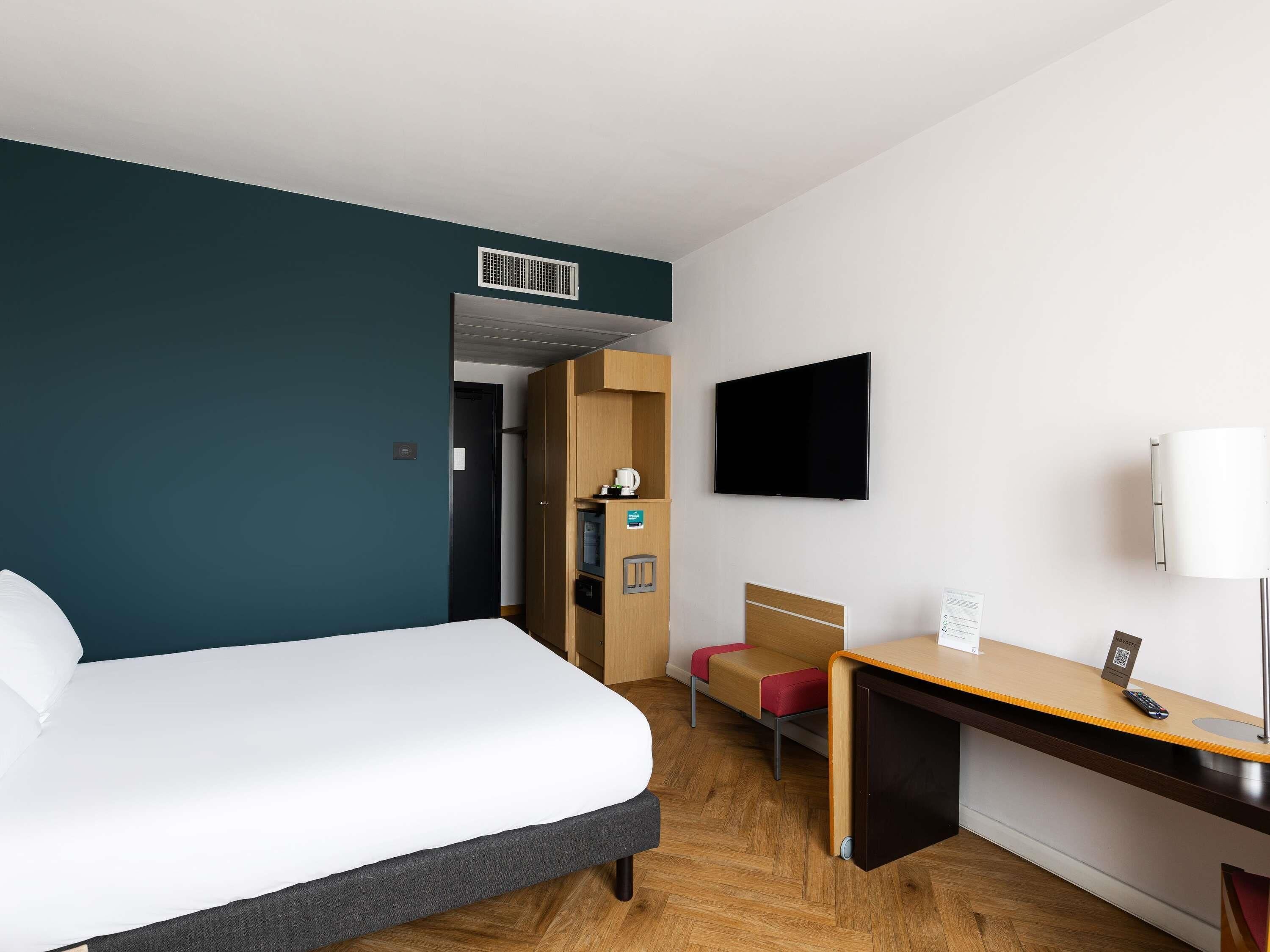 Quarto de hotel com cama, escrivaninha, cadeira, televisão e minibar.