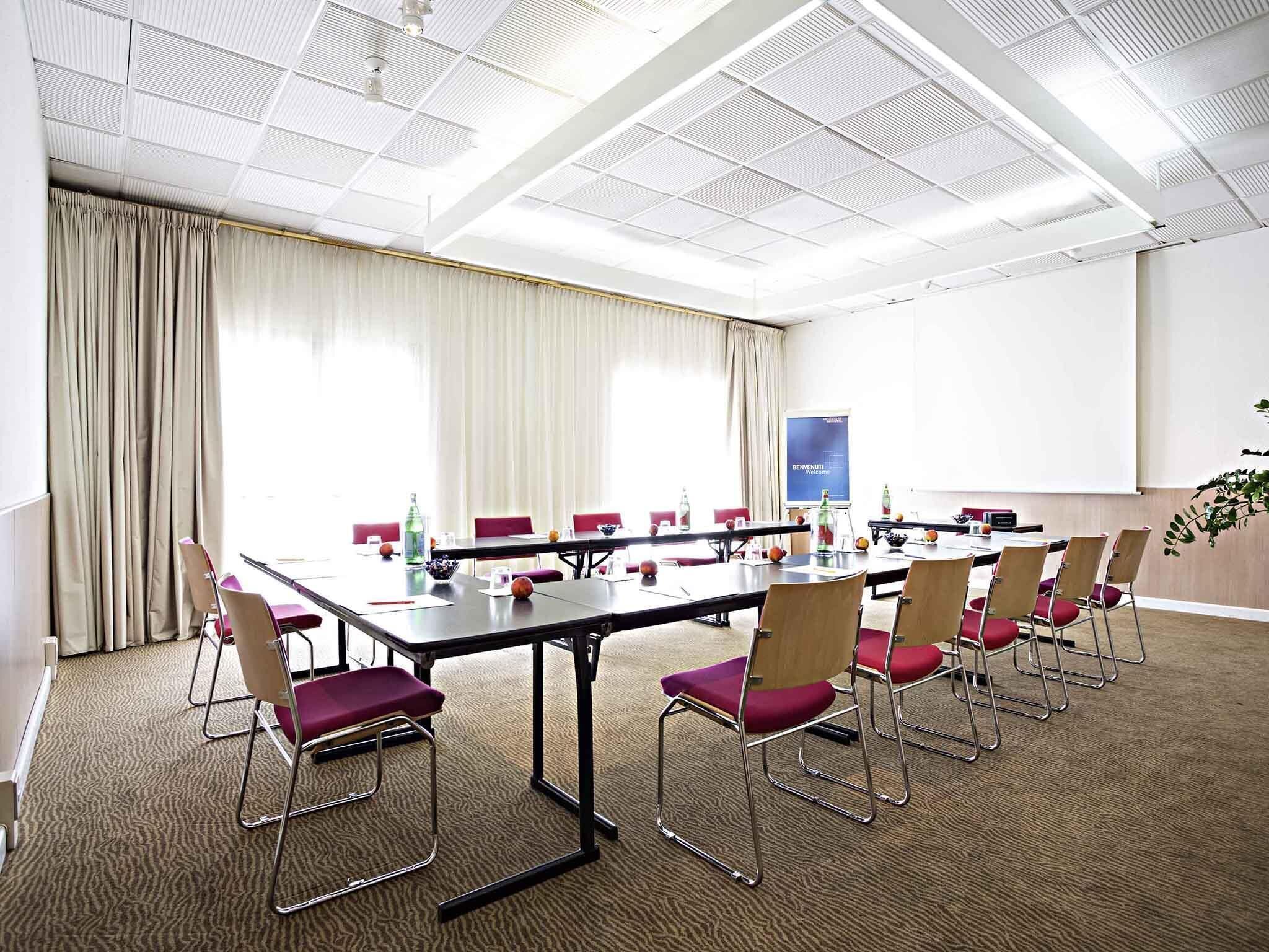 Une salle de conférence avec une longue table, des chaises rouges, un écran de projection et une grande fenêtre avec des rideaux.