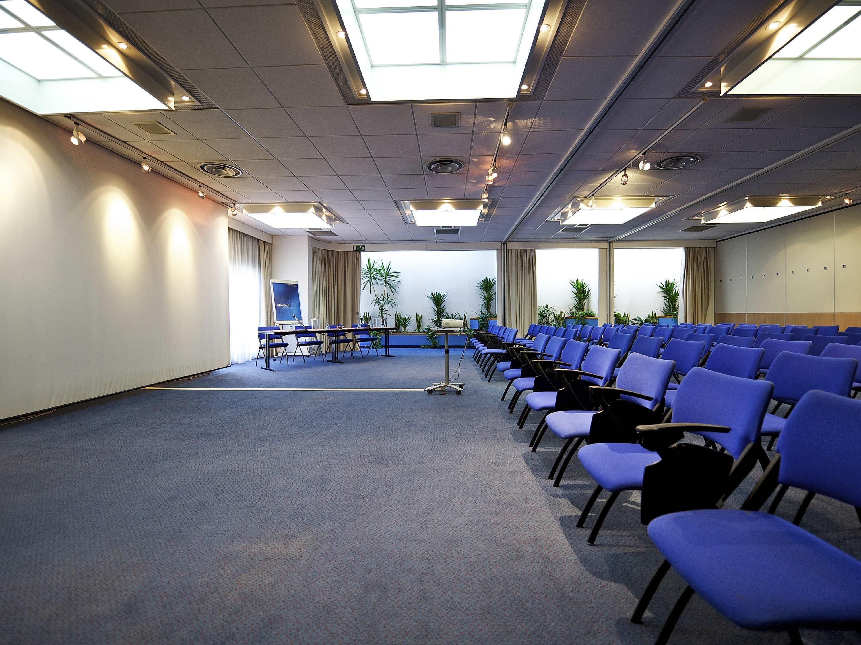 Une salle de conférence avec des rangées de chaises bleues, un écran de présentation et des plantes en pot.