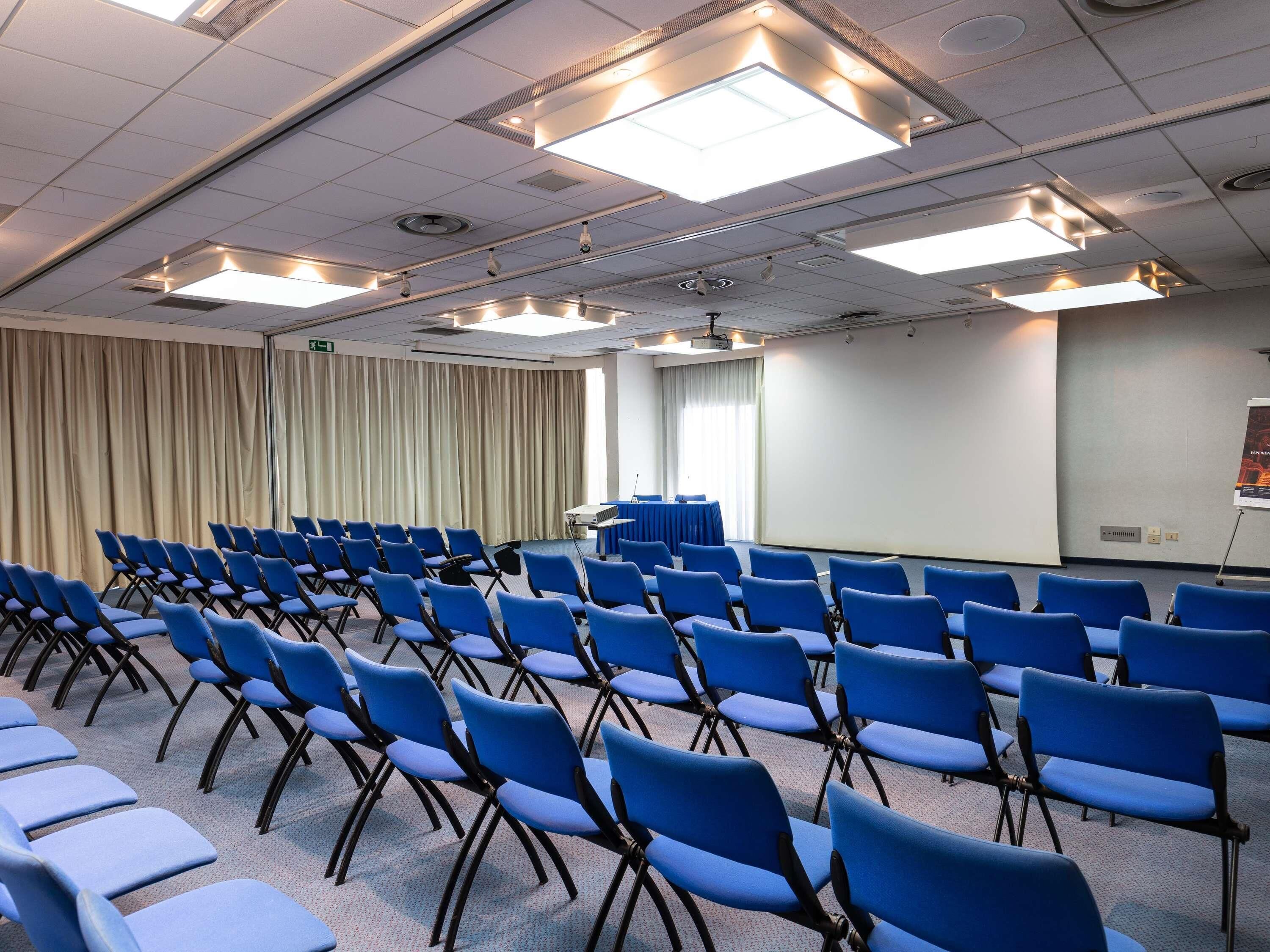 Ein Konferenzraum mit Reihen blauer Stühle, einer Projektionsleinwand und Deckenleuchten.