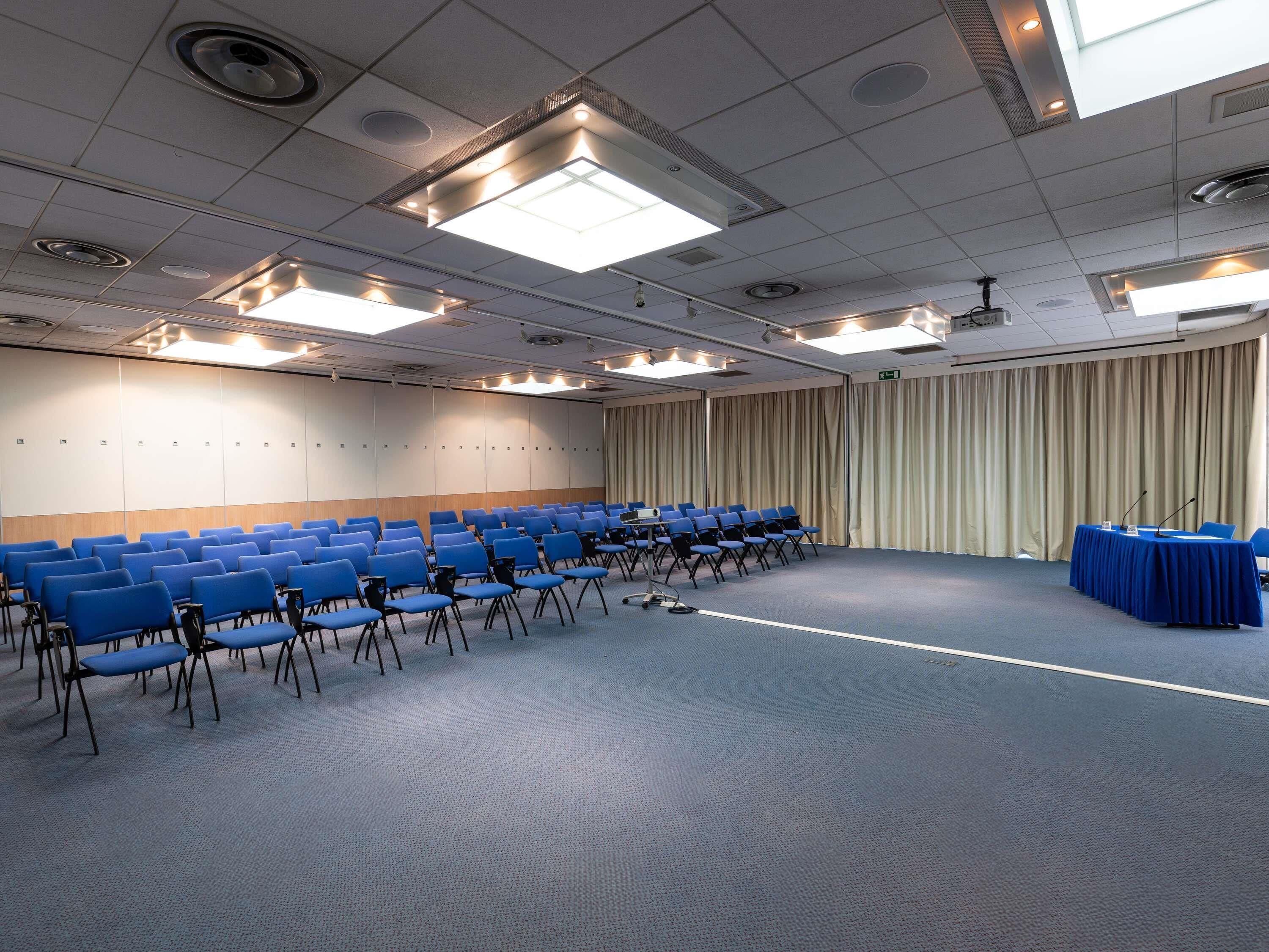 Ein Konferenzraum mit blauen Stühlen, einer Bühne mit einem blauen Tisch und einem Vorhang im Hintergrund.