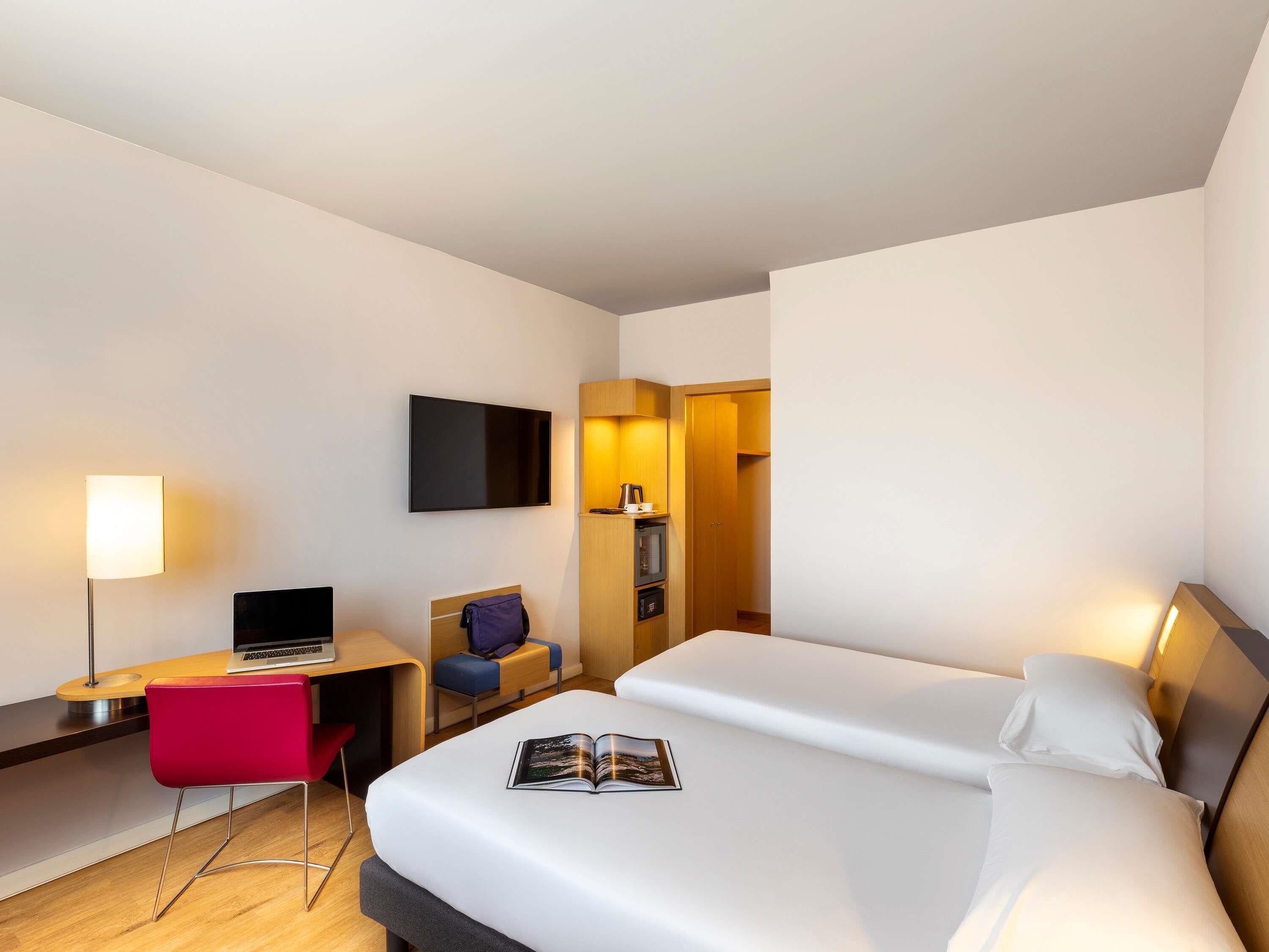 Una habitación de hotel con cama, escritorio con una computadora portátil, silla, televisión y mesita de noche con lámpara.