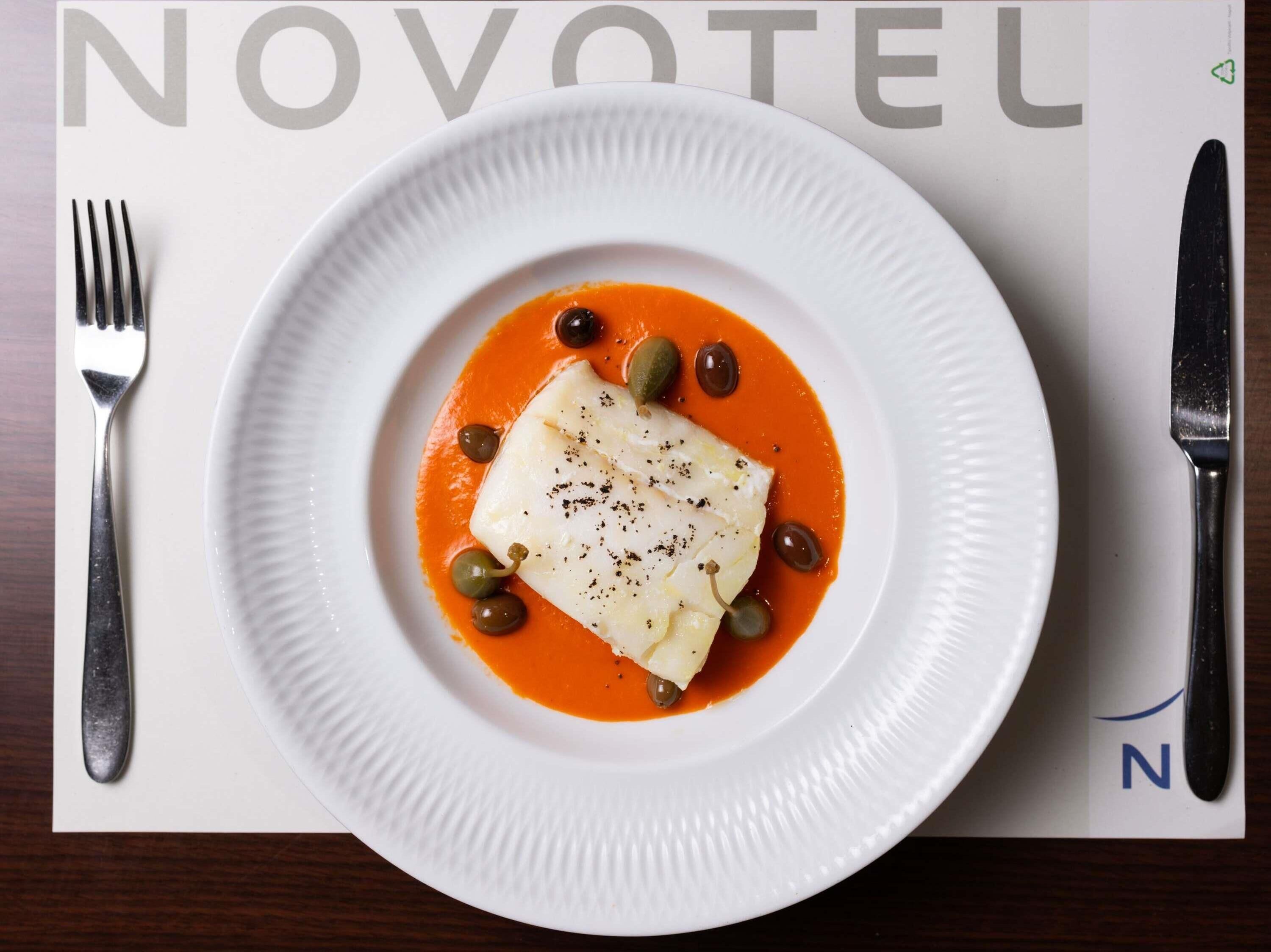 A white plate with a white fish fillet, black pepper, and green olives on a white plate, set on a table with a fork and knife.