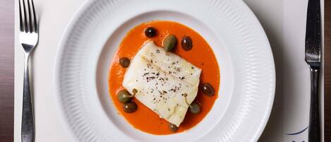 A white plate with a white fish fillet, black pepper, and green olives on a white plate, set on a table with a fork and knife.