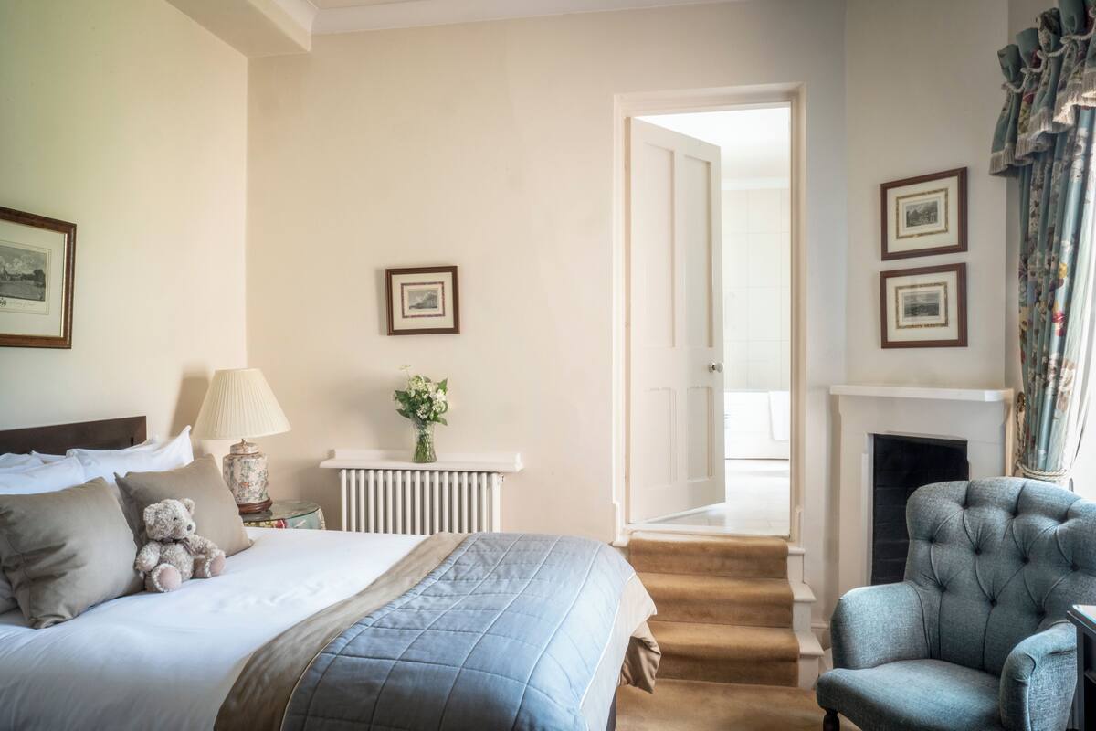 A bedroom with a bed, a chair, a lamp, a fireplace, and framed pictures on the wall.