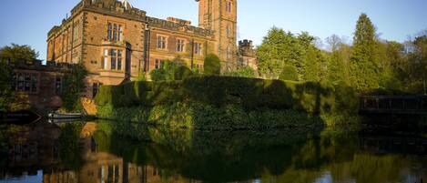 A historic building with a tower, reflecting in a calm body of water, surrounded by greenery.