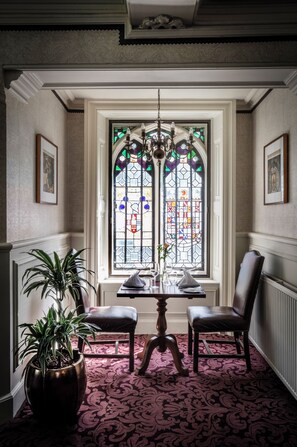 A dining area with a table and chairs, a chandelier, a stained glass window, and a potted plant. - New Hall Hotel & Spa (Sutton Coldfield)