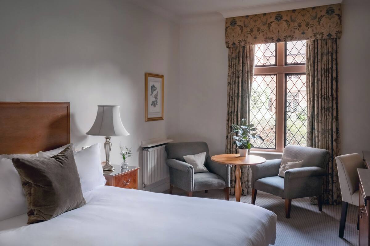 A hotel room with a bed, two armchairs, a small table, a lamp, a radiator, and a window with curtains.