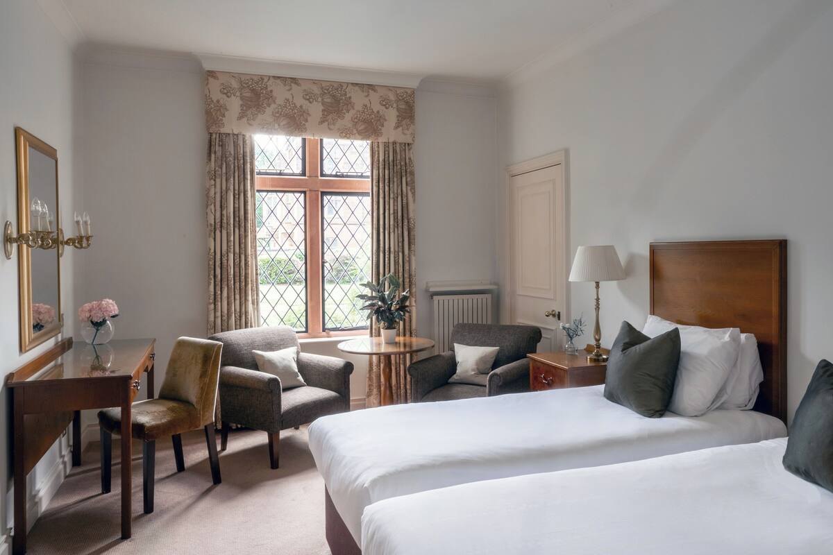 A hotel room with two beds, a desk, a chair, a table with a plant, and a window with curtains.