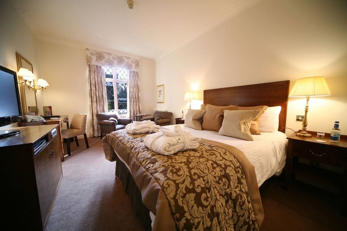A hotel room with a bed, two chairs, a desk, a television, and a window with curtains.
