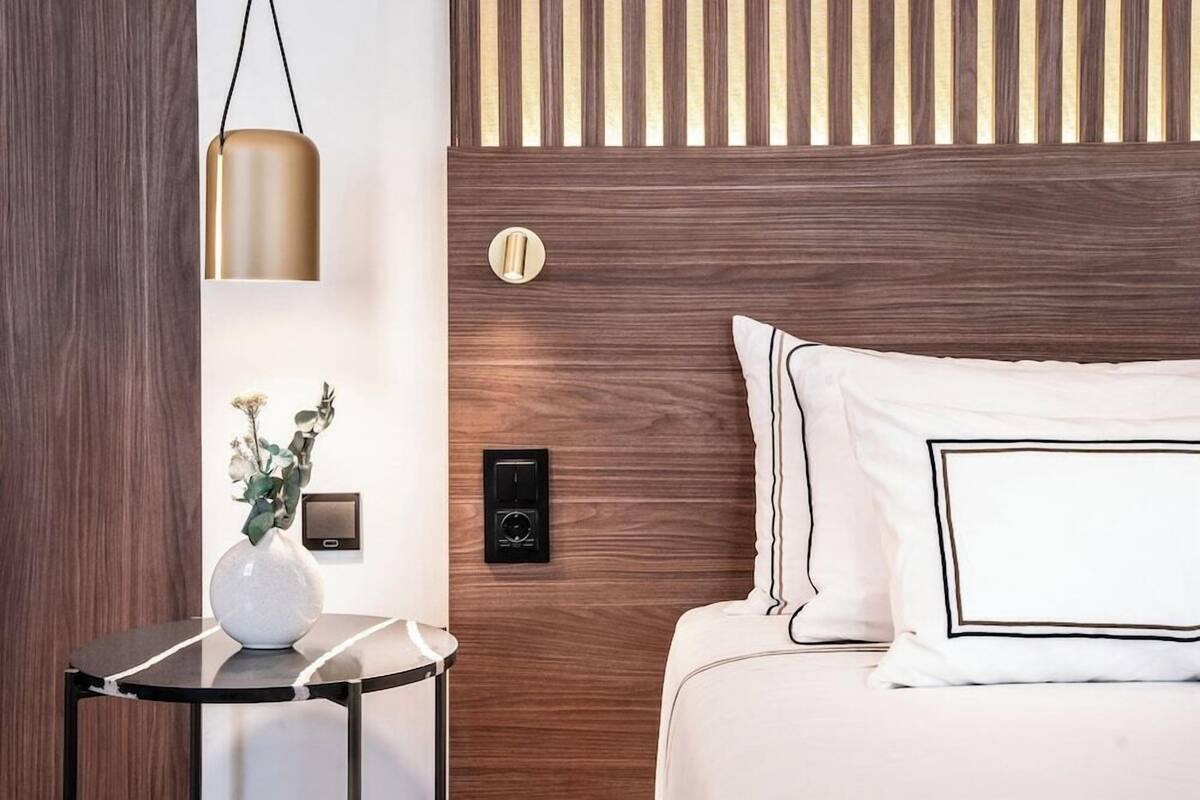 Una habitación de hotel con cabecera de madera, una mesita de noche con un florero y un panel de control en la pared.