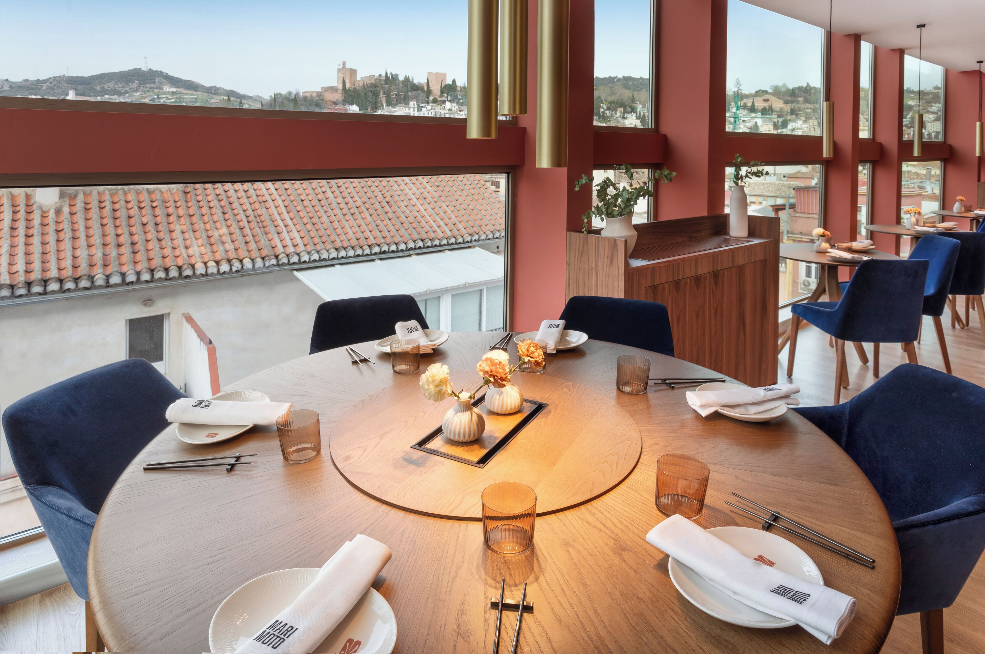 A dining area with a round table, blue chairs, and a view of the cityscape through large windows.