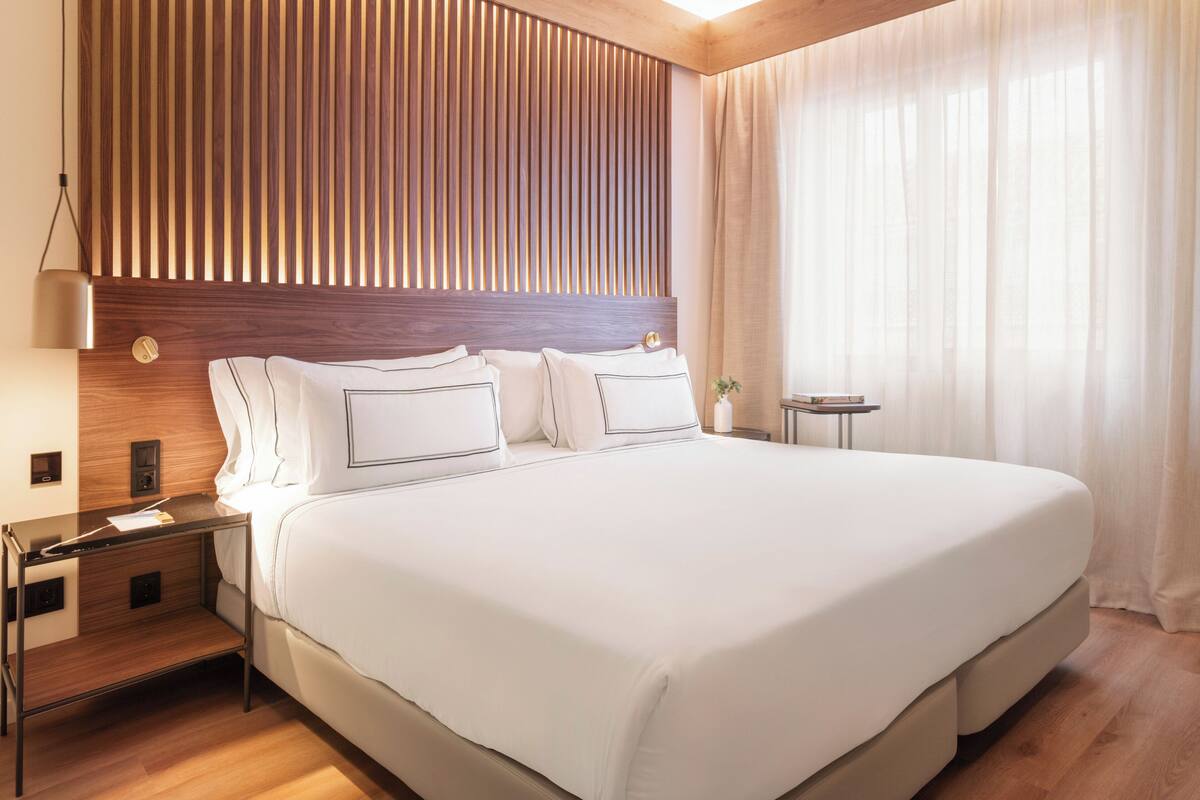 Una cama grande con ropa blanca, un cabecero de madera, una mesita de noche con lámpara y un pequeño atril.