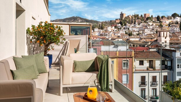 A balcony with a sofa, a coffee table with a glass and a bottle, and a view of a cityscape with buildings and a hill. - Melia Granada Hotel (Granada)
